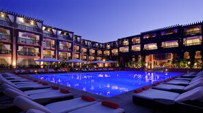 Un hotel con zona de piscina que cuenta con tumbonas y sombrillas, un edificio de varios pisos con balcones y una fachada bien iluminada.