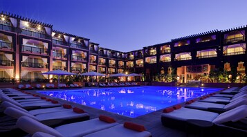 A hotel with a poolside area featuring sun loungers and umbrellas, a multi-story building with balconies, and a well-lit facade.
