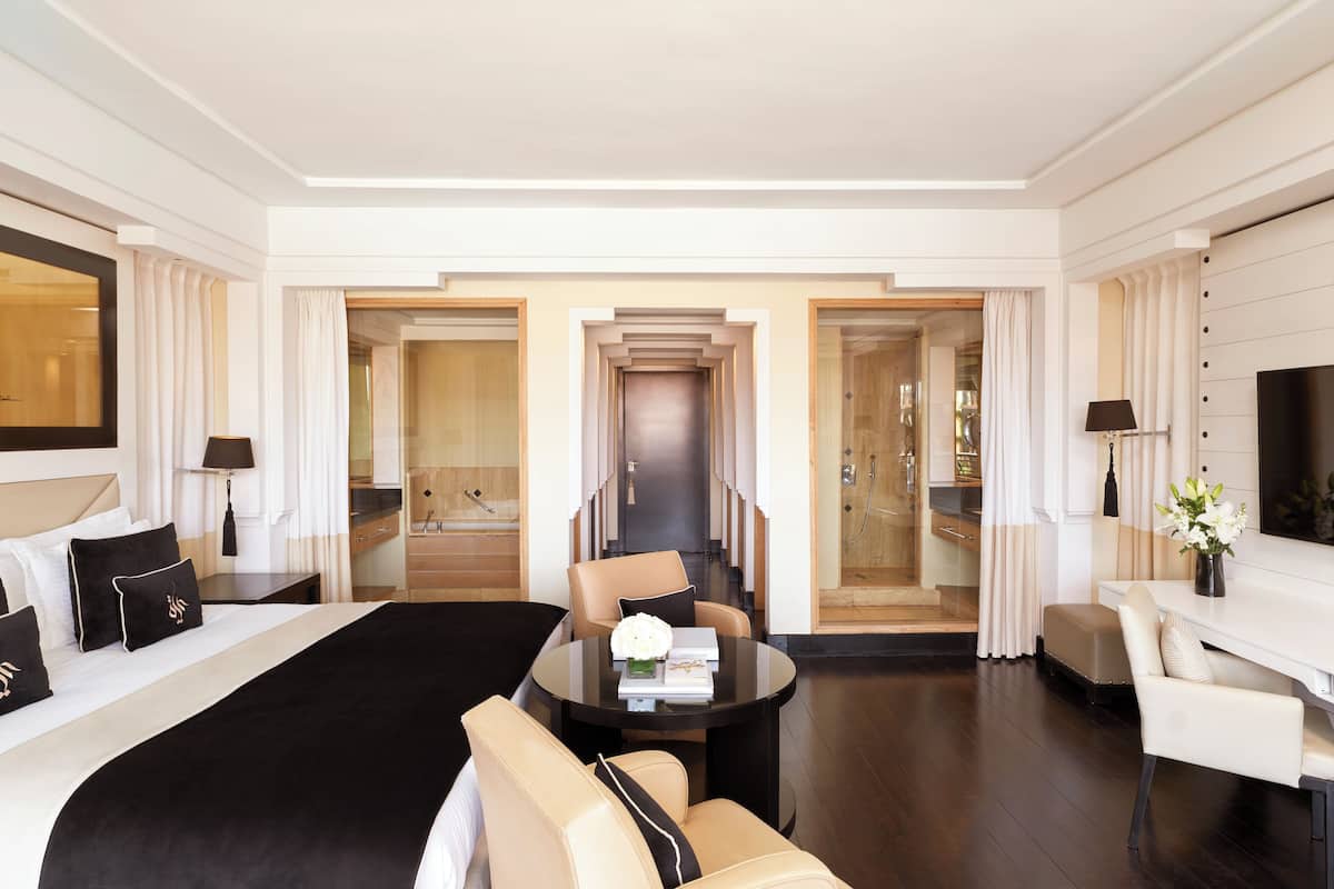 A hotel room with a large bed, a sitting area with a table and chairs, a TV, and a bathroom visible through an open door.
