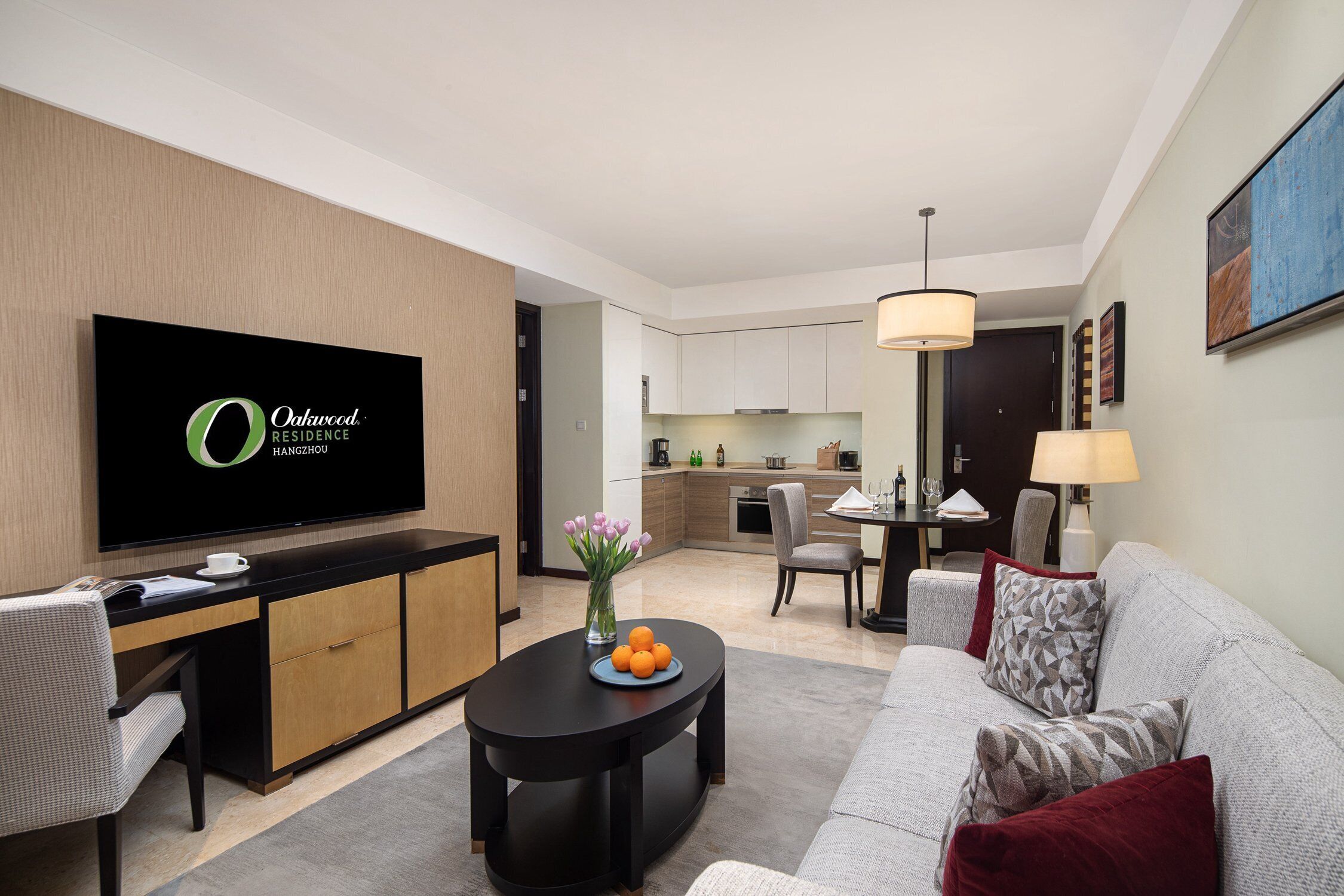 A modern living room with a flat-screen TV, a dining table with chairs, a sofa with cushions, and a kitchen in the background.