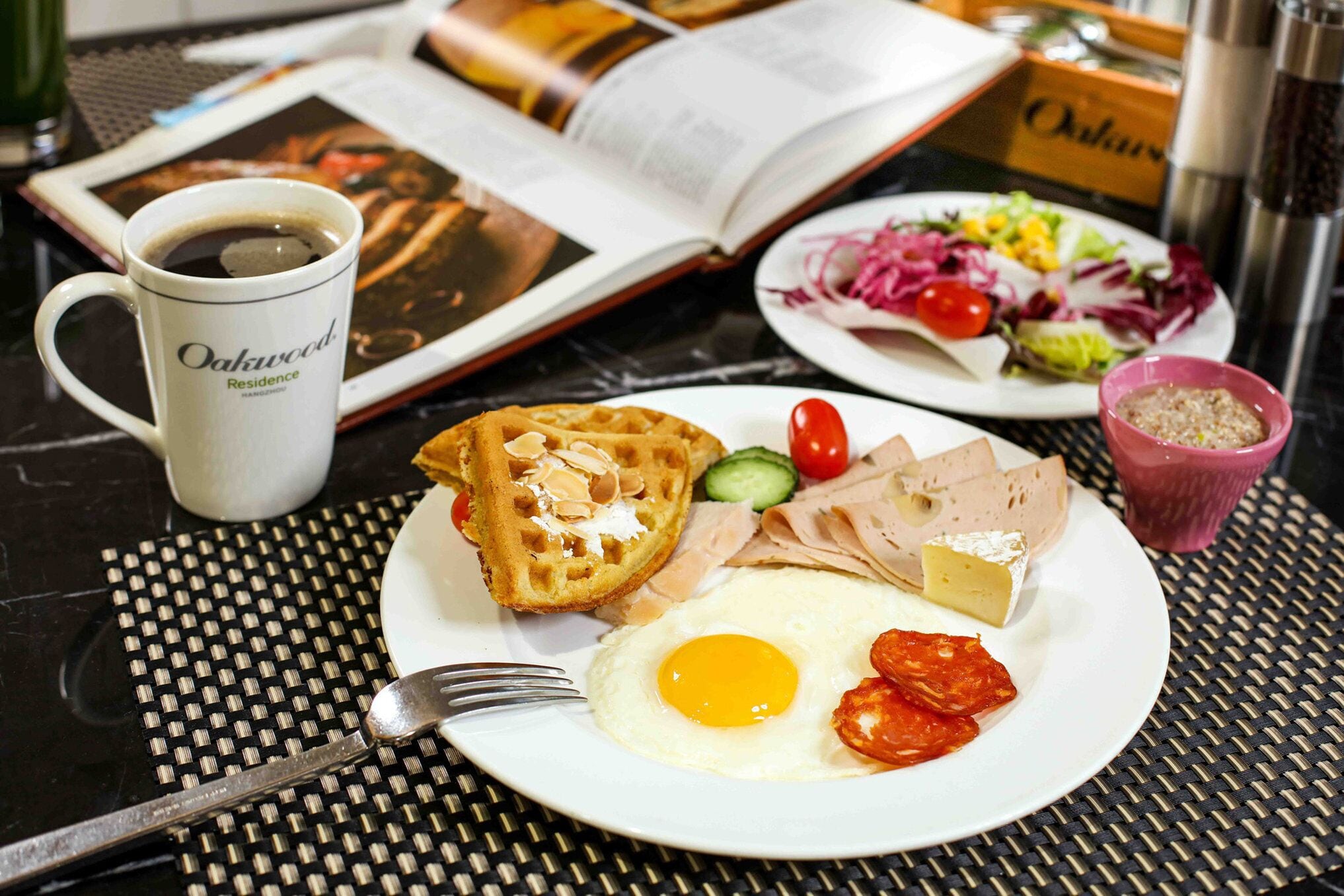 A breakfast plate with waffles, a fried egg, and ham, a cup of coffee, and a salad on a table with a book and a magazine.