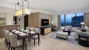A modern hotel room with a dining area, a living room with a sofa and armchairs, a TV, and a city view.