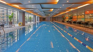 An indoor swimming pool with clear lanes, a sun terrace with seating, and large windows offering views of the surrounding buildings.