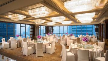 Un amplio salón de banquetes con varias mesas redondas, elegantes candelabros y grandes ventanales con vistas a la ciudad.