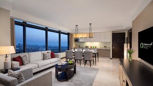 A modern living room with a sofa, coffee table, and dining area. A large TV is mounted on the wall, and there is a view of the city through large windows.