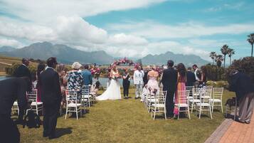 An outdoor wedding ceremony with guests seated on white chairs arranged in rows, facing the couple standing under a floral arch.