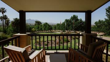 A balcony with wooden chairs and a table, overlooking a landscaped garden with trees and a distant mountain range.
