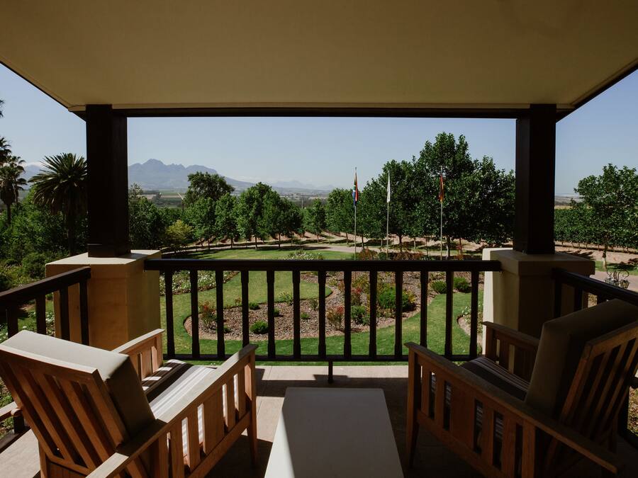 A balcony with wooden chairs and a table, overlooking a landscaped garden with trees and a distant mountain range.