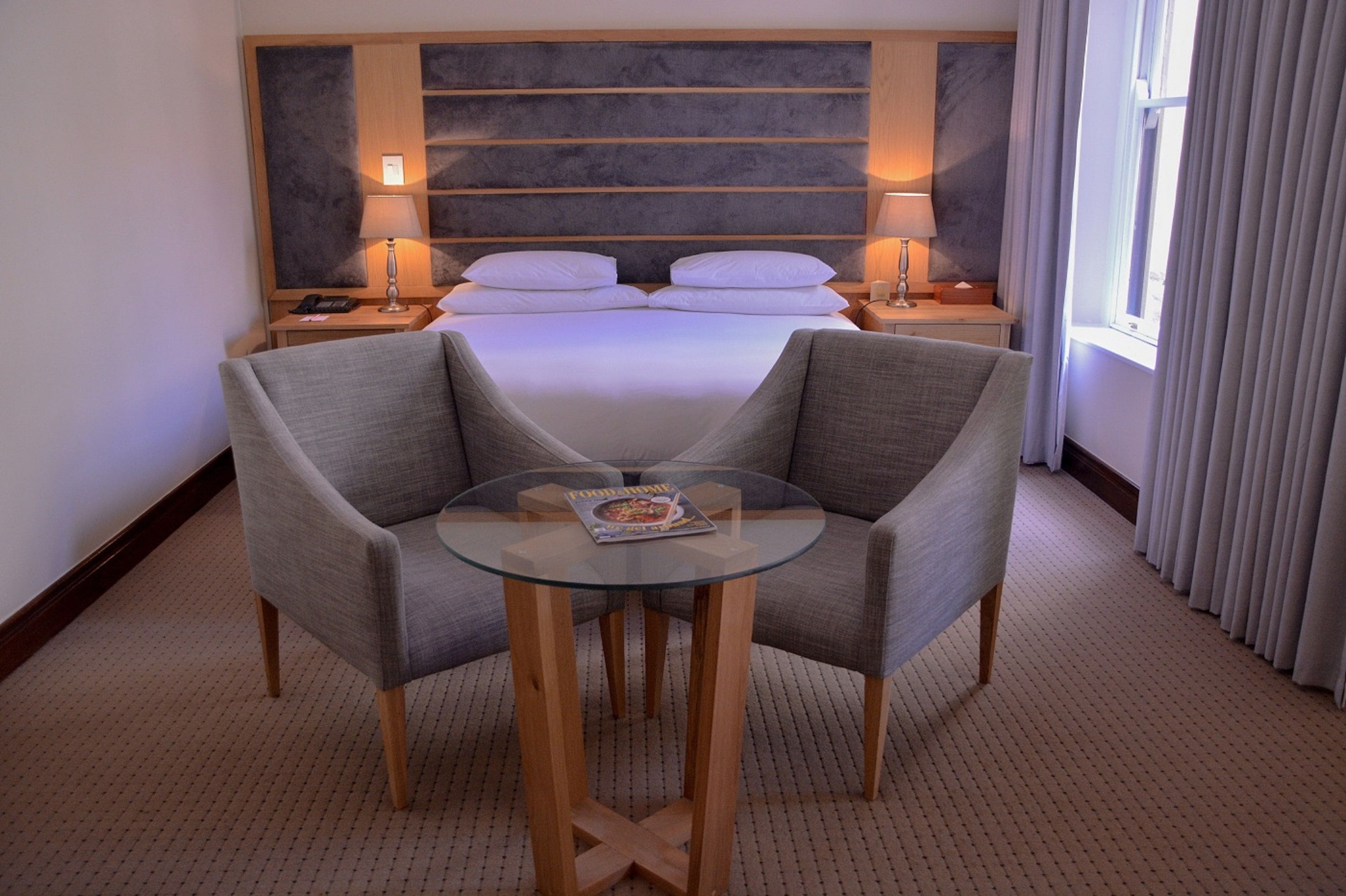 A hotel room with a large bed, two bedside tables with lamps, a glass table with two chairs, and a view of the outside through a window.