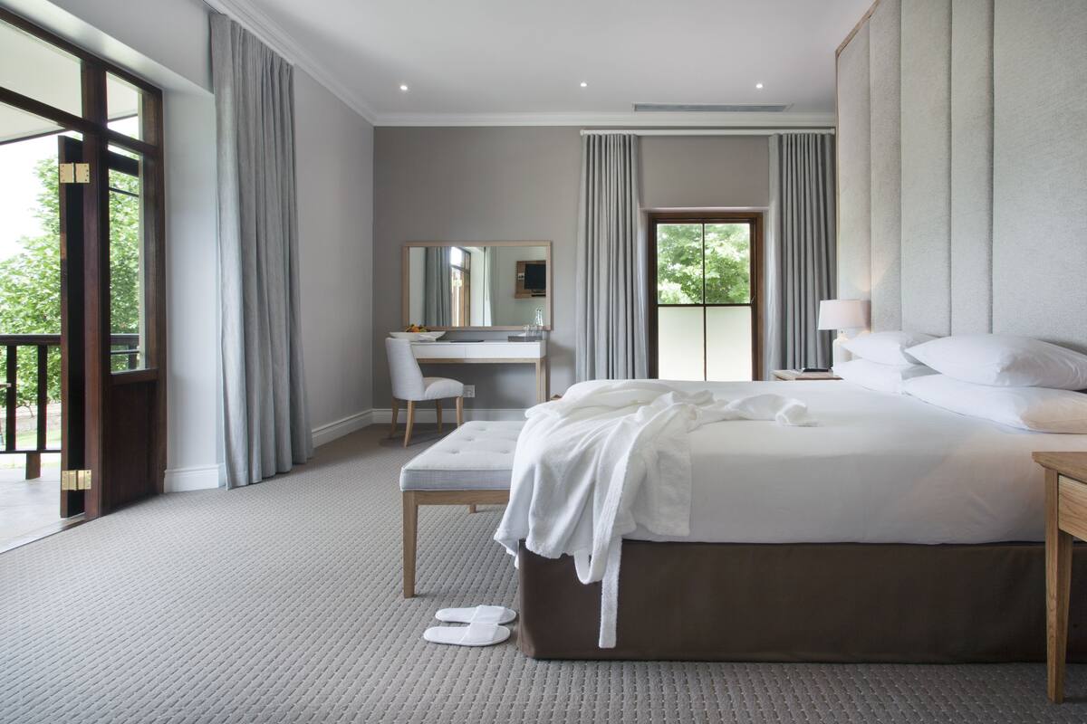 A modern bedroom with a large bed, a desk, and a chair. There is a window with curtains and a view of greenery outside.