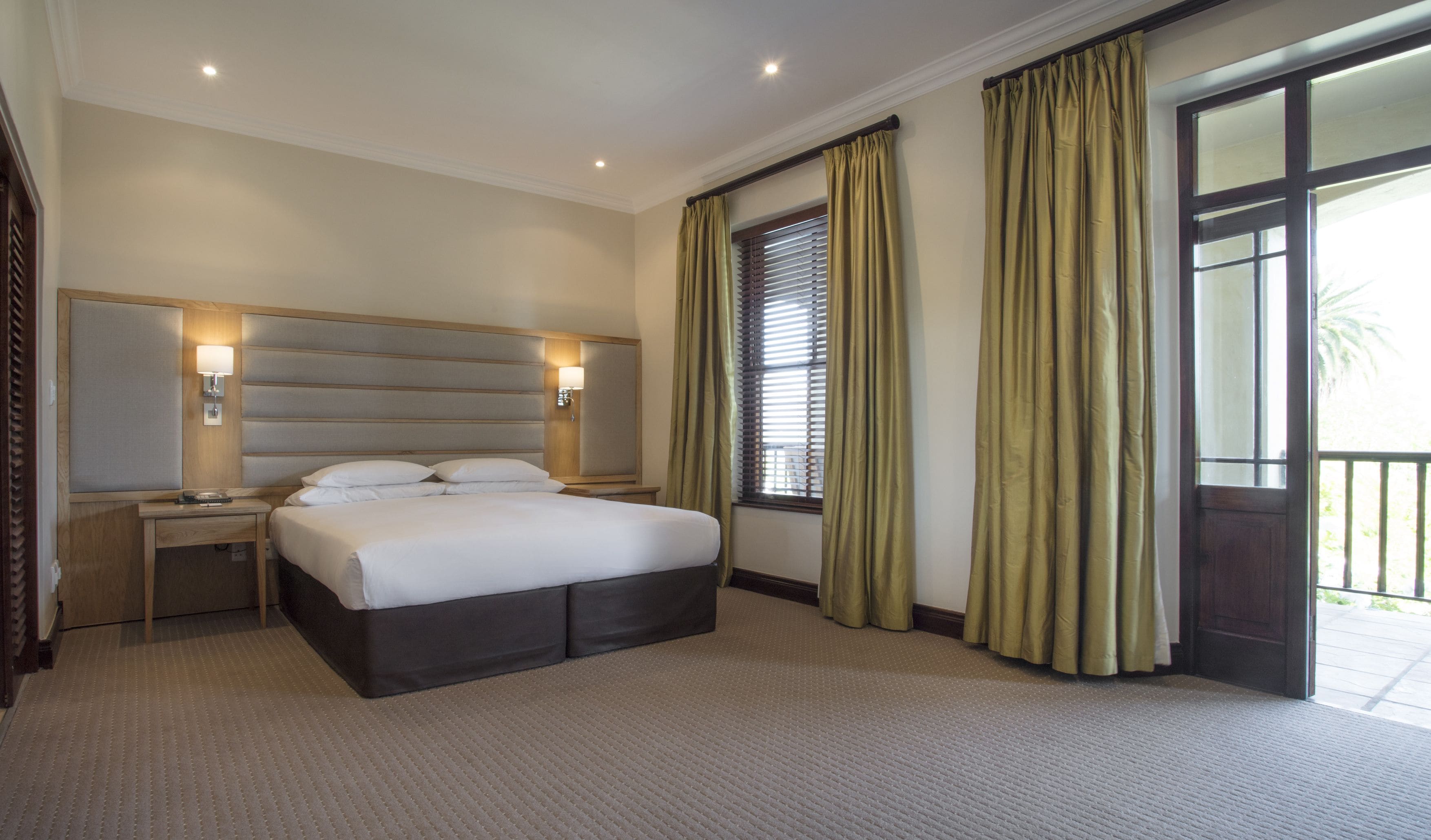 Une chambre d’hôtel avec un grand lit, deux tables de chevet, une tête de lit, une fenêtre avec des stores et une porte donnant sur un balcon.