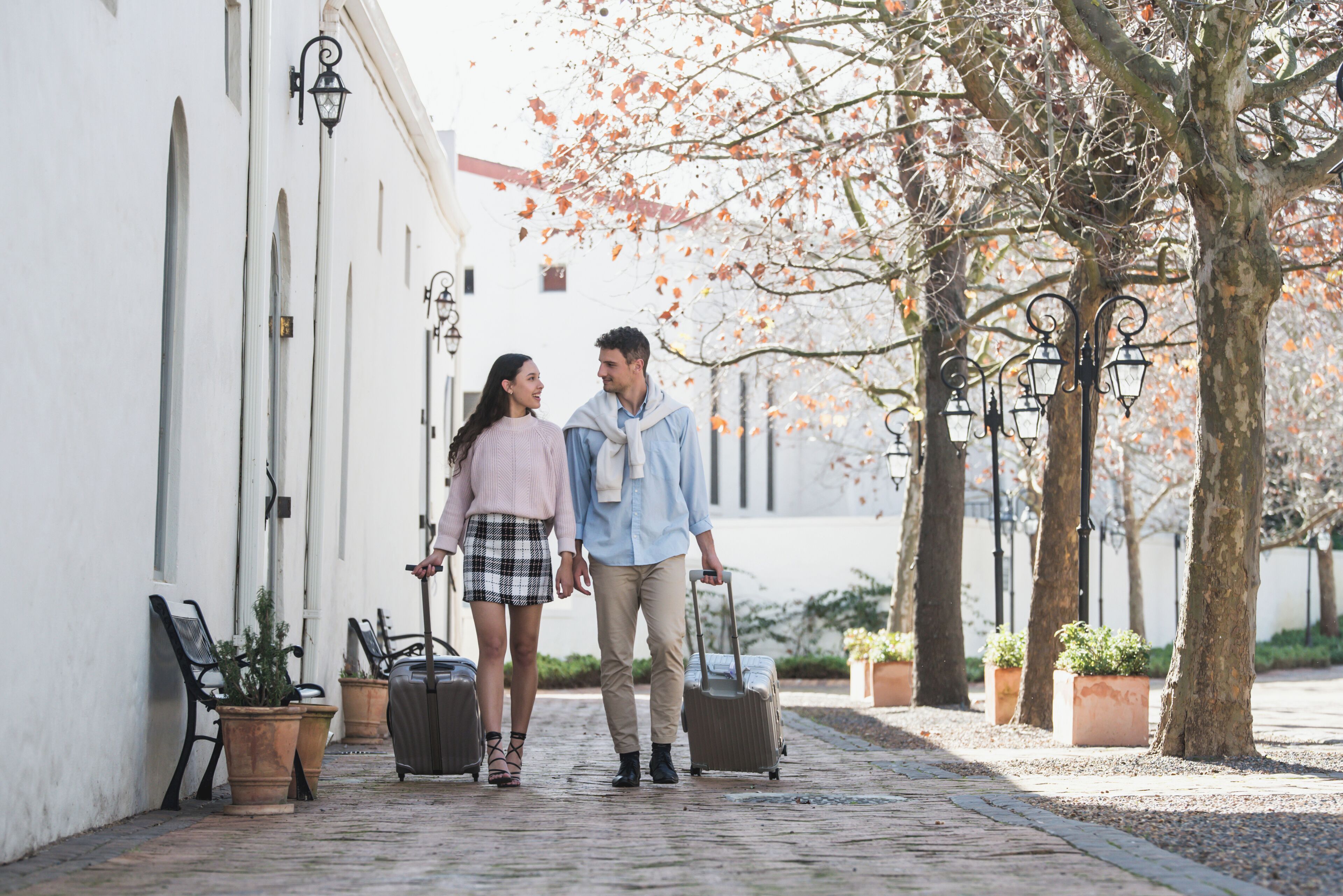 A couple walking on a cobblestone path with a suitcase, flanked by white buildings and trees with autumn leaves.