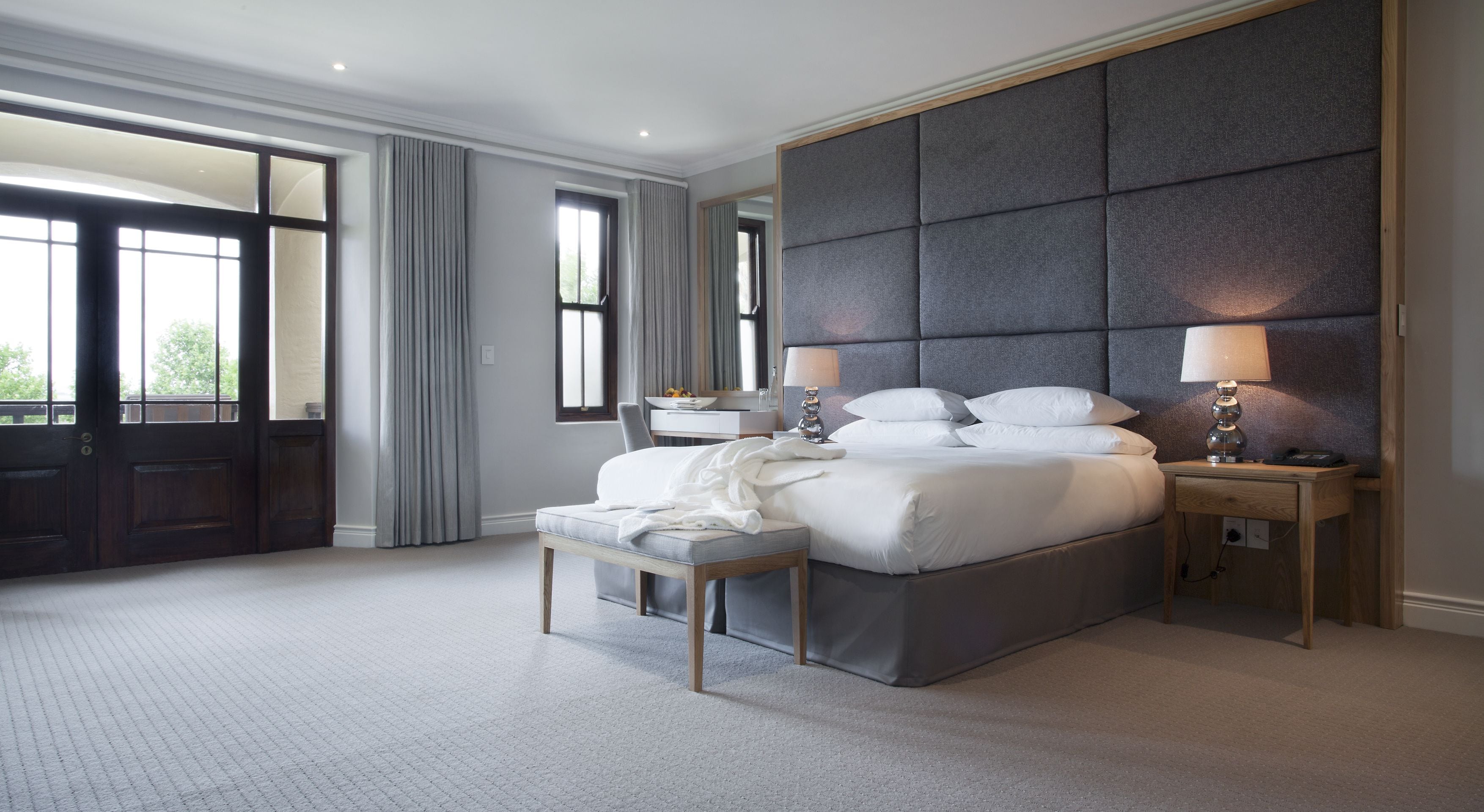 A modern bedroom with a large bed, a bedside table with lamps, a chair, and a desk with a mirror.