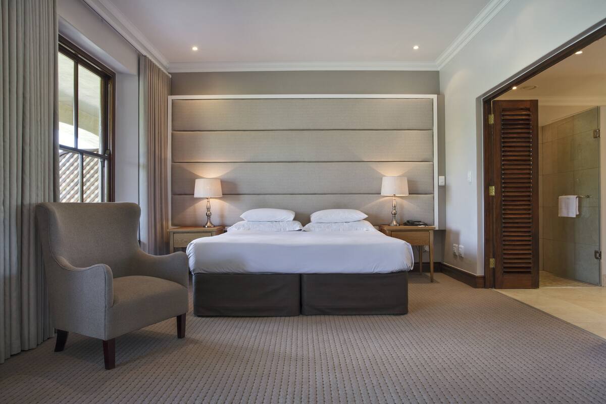 A hotel room with a large bed, a chair, two bedside tables with lamps, and a door leading to a bathroom.