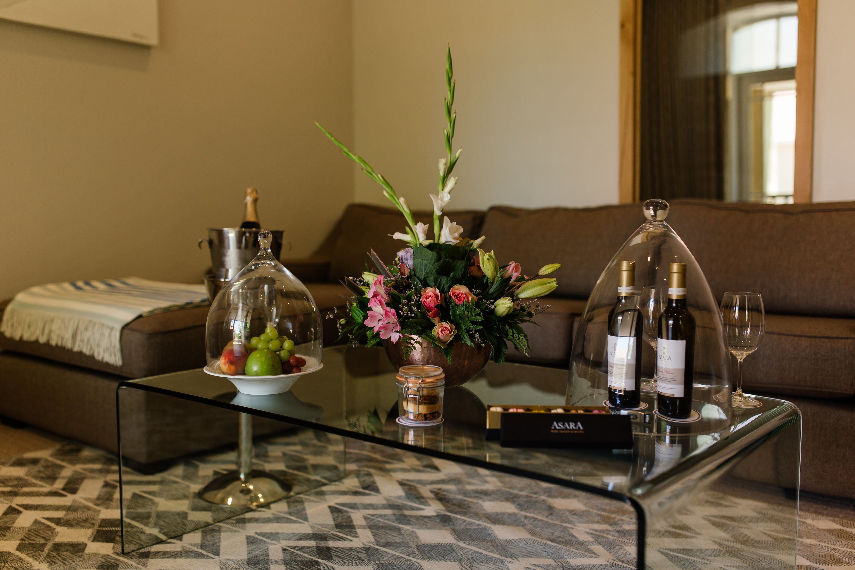 A glass coffee table with a bouquet of flowers, a champagne bottle, and two wine bottles in front of a brown sofa.