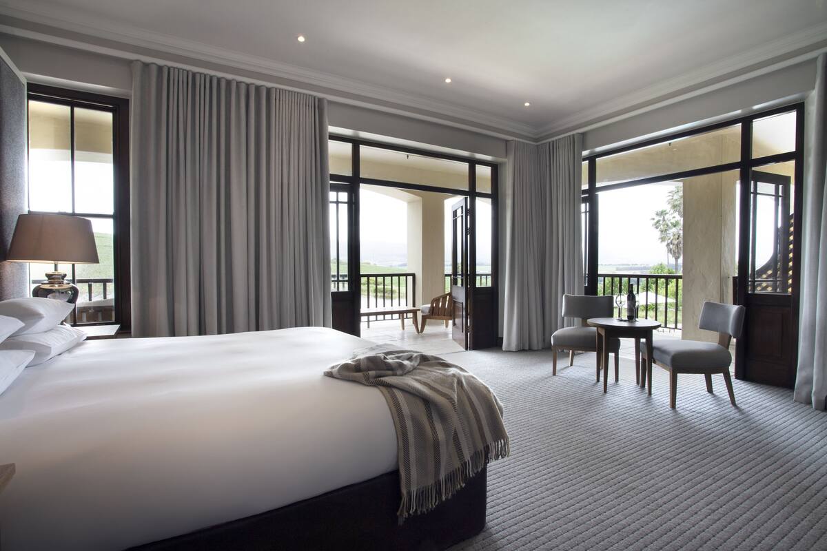 A hotel room with a large bed, a desk with two chairs, and a balcony with a view.