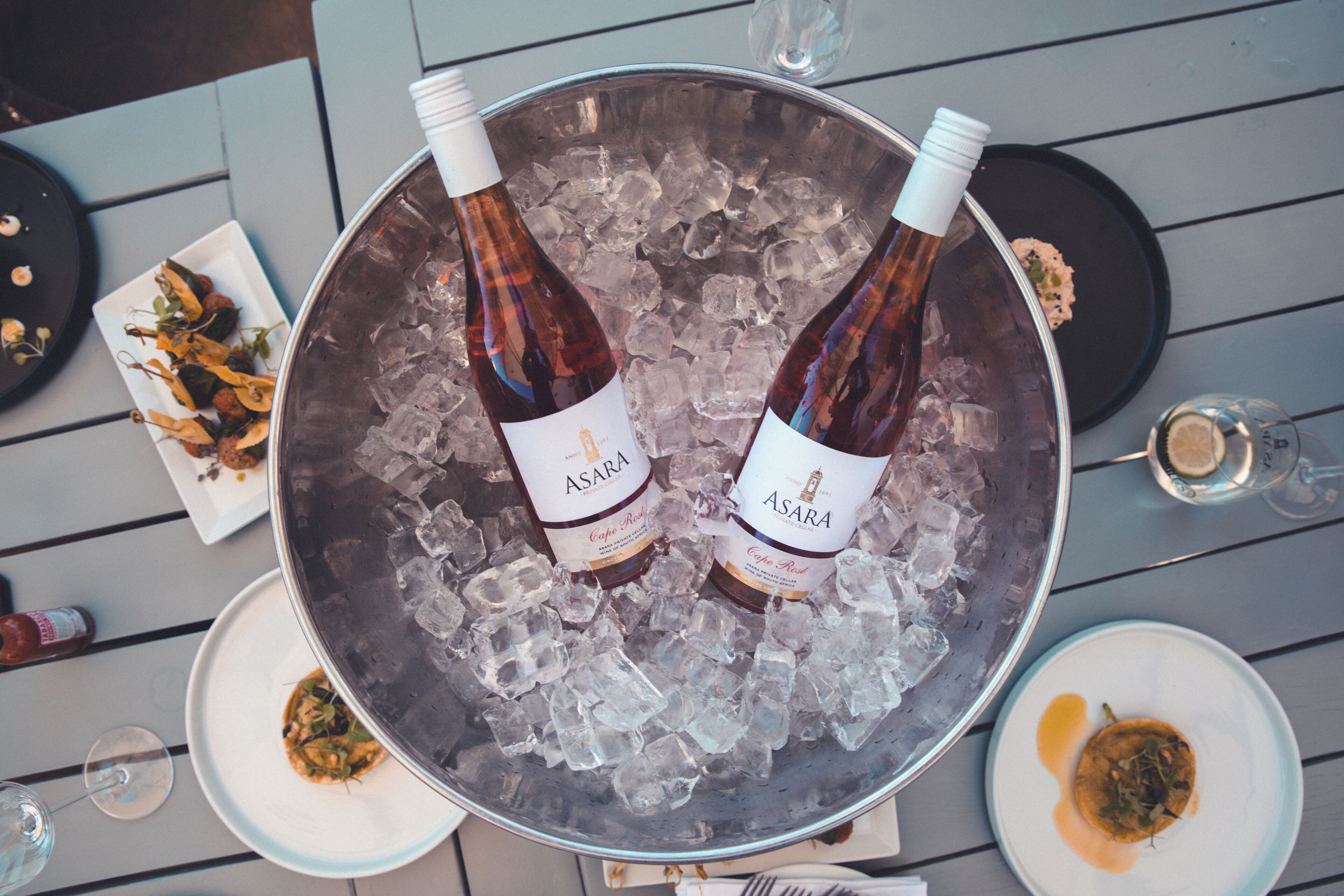 Dos botellas de vino Asara en un balde de hielo sobre una mesa con platos de comida.