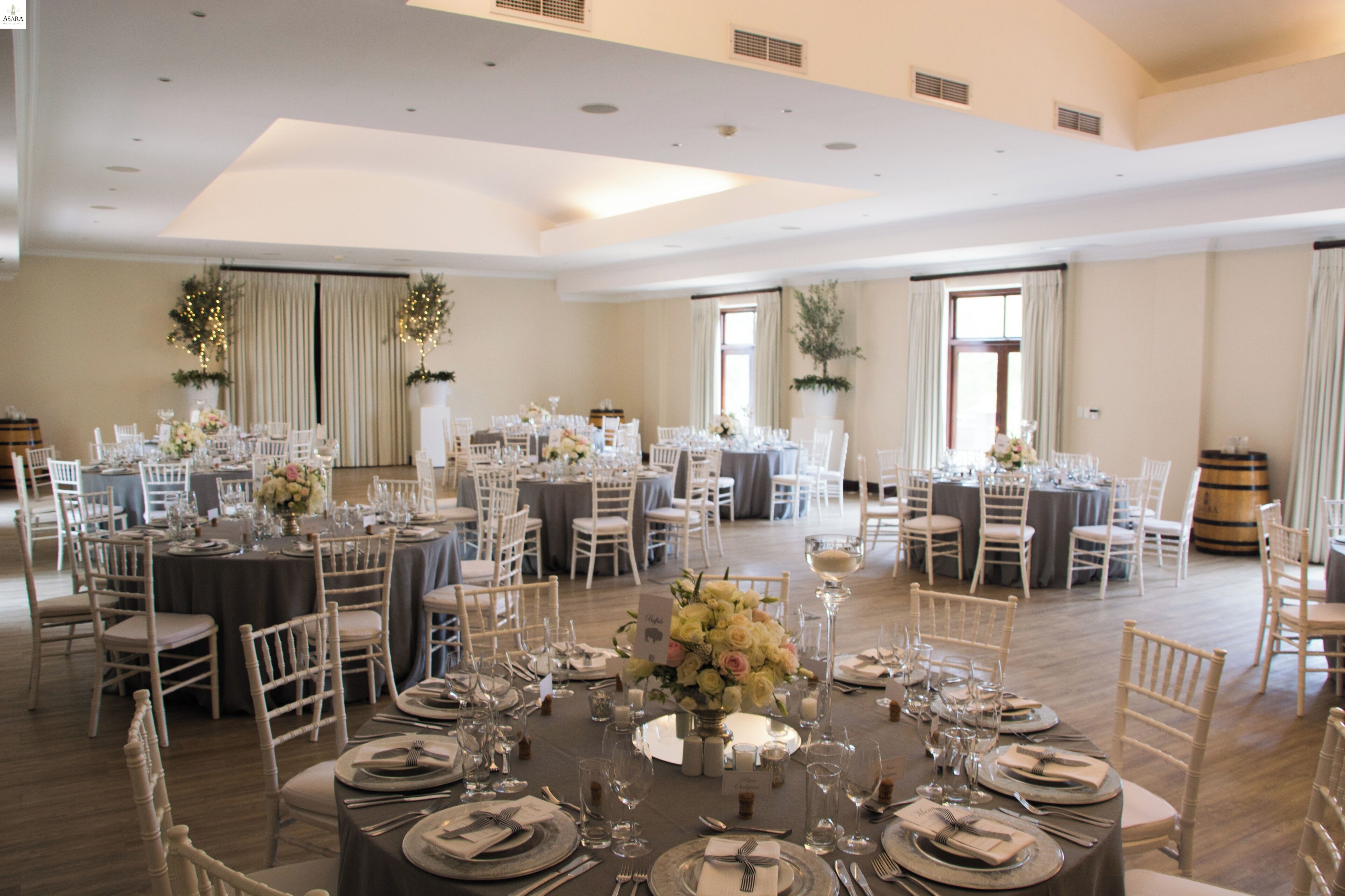 A spacious banquet hall with multiple round tables set for dining, surrounded by white chairs.