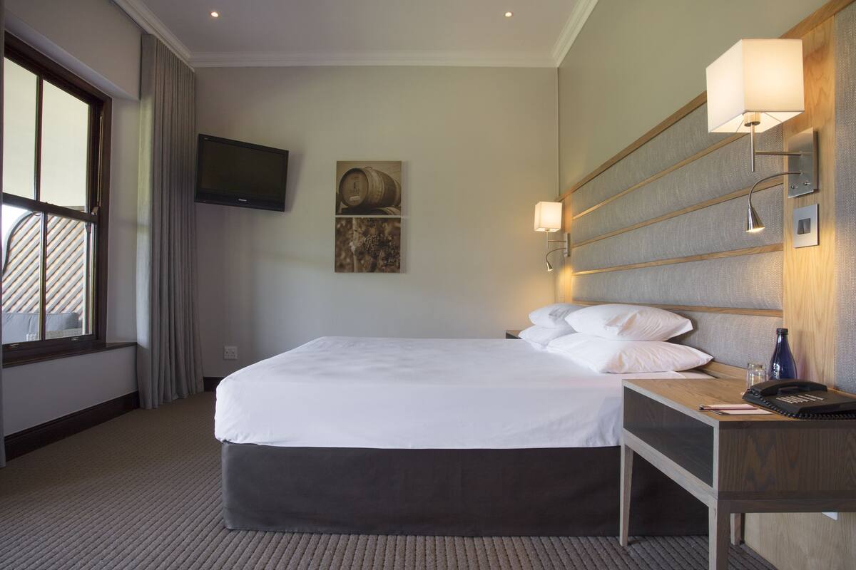 A hotel room with a large bed, a bedside table with a lamp, a flat-screen TV mounted on the wall, and a window with curtains.