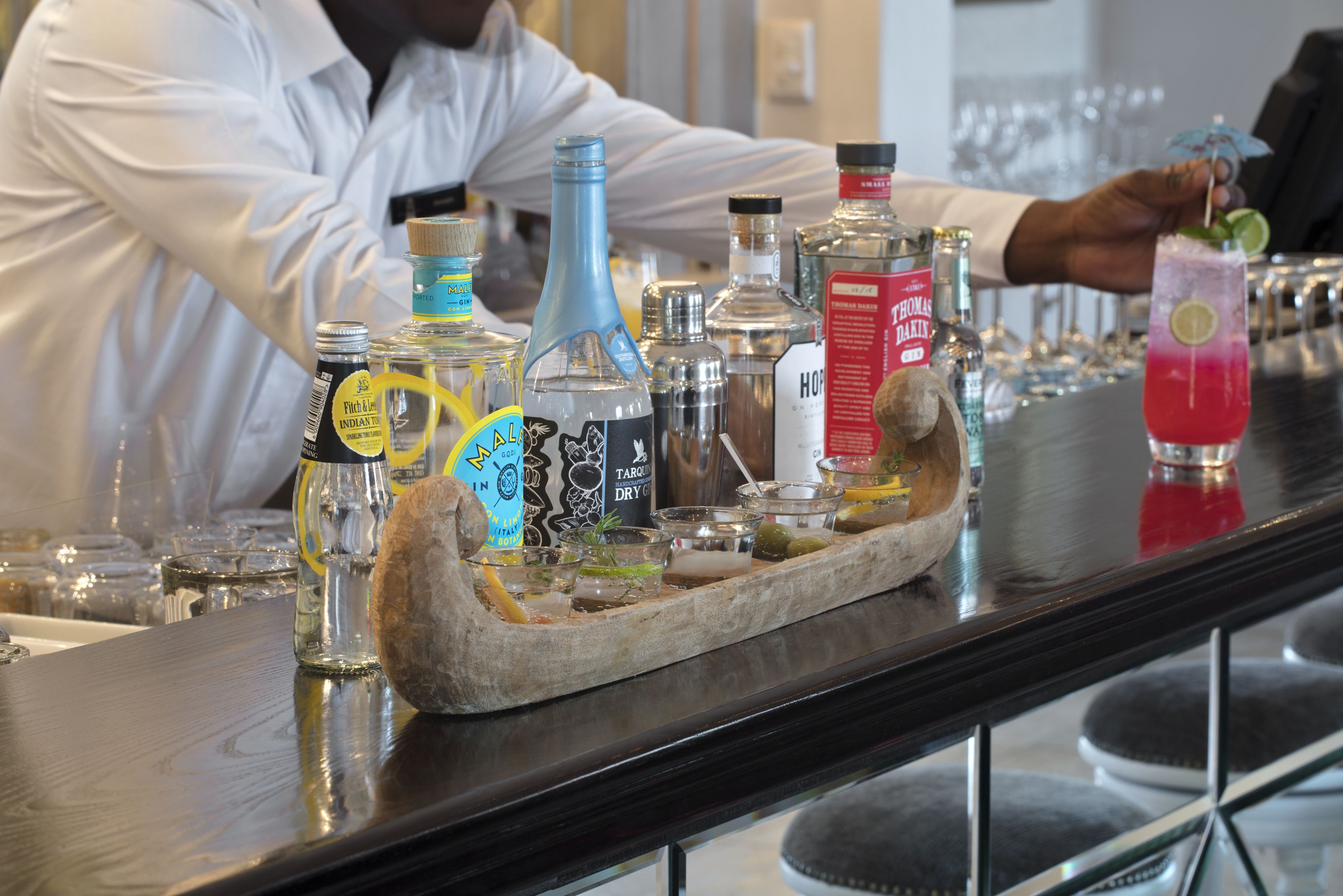 Un coin bar avec différentes bouteilles et différents verres, un plateau en bois et une personne qui prépare une boisson.