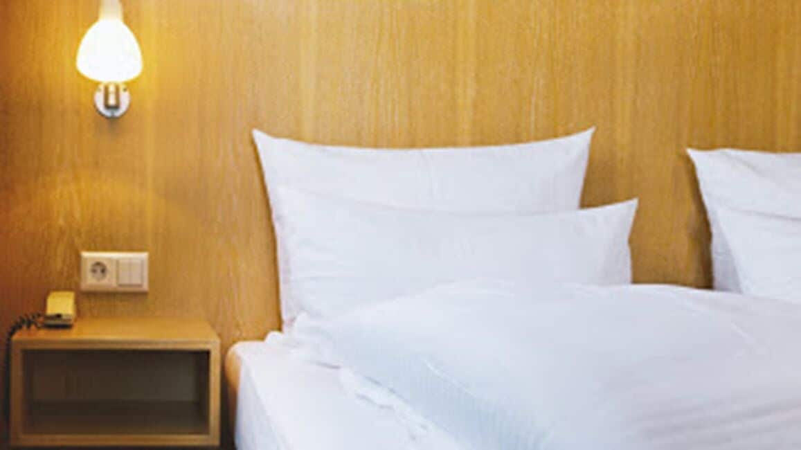 A hotel room with a wooden headboard, a bedside table with a lamp, and a bed with white bedding.