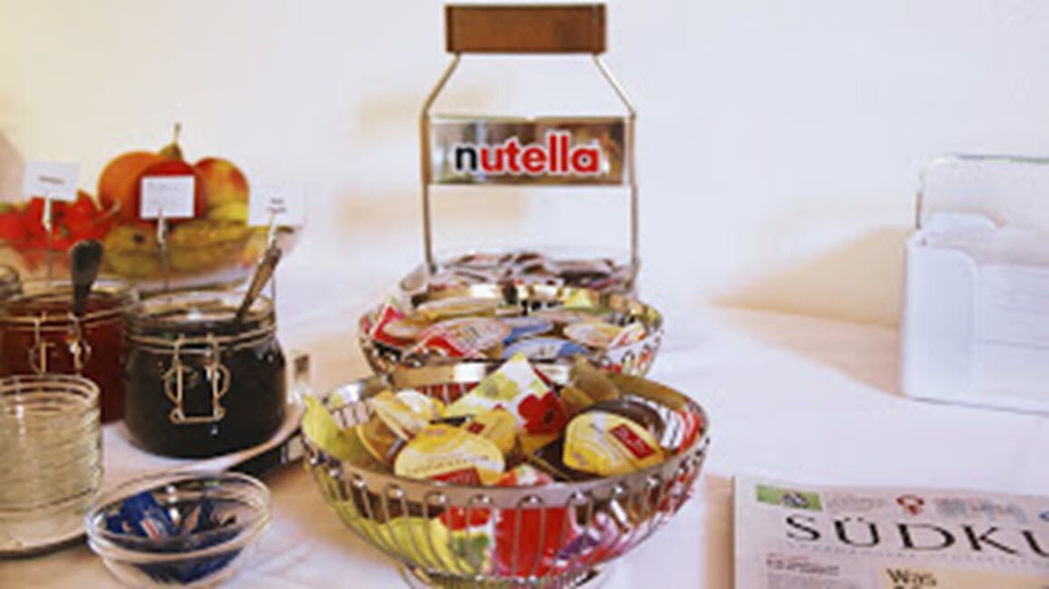 Una mesa de buffet con diferentes alimentos, incluyendo frascos de mermelada, un tazón de fruta y un recipiente con 'Nutella'.