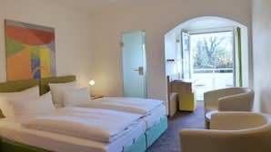 A hotel room with a double bed, a desk with a chair, a small table, and a view through an open door. - Hotel Alpenblick Garni (Ueberlingen)