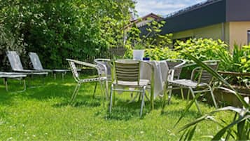A grassy outdoor area with several white metal chairs and a table, surrounded by greenery and a building in the background.