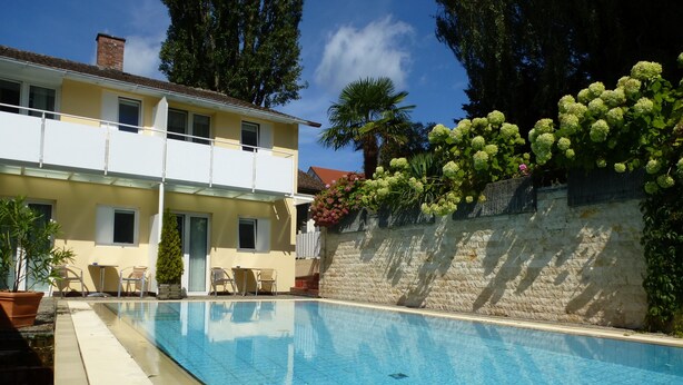 A swimming pool with a clear blue water surface, surrounded by a stone wall and a yellow building with windows and doors. - Hotel Alpenblick Garni (Ueberlingen)
