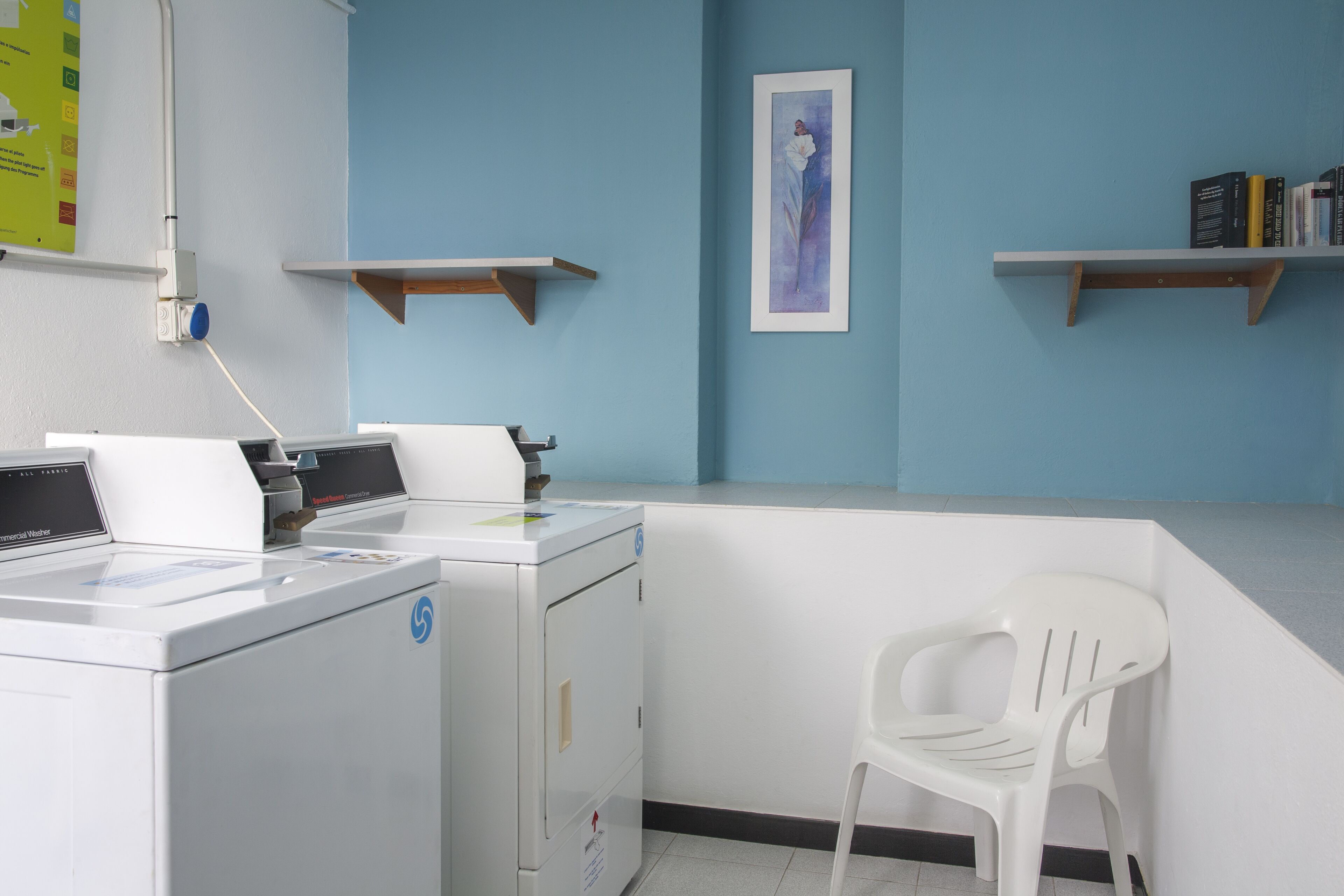 A laundry room with a washing machine, dryer, and a white plastic chair. There are two wooden shelves against the wall, one with books and the other empty.