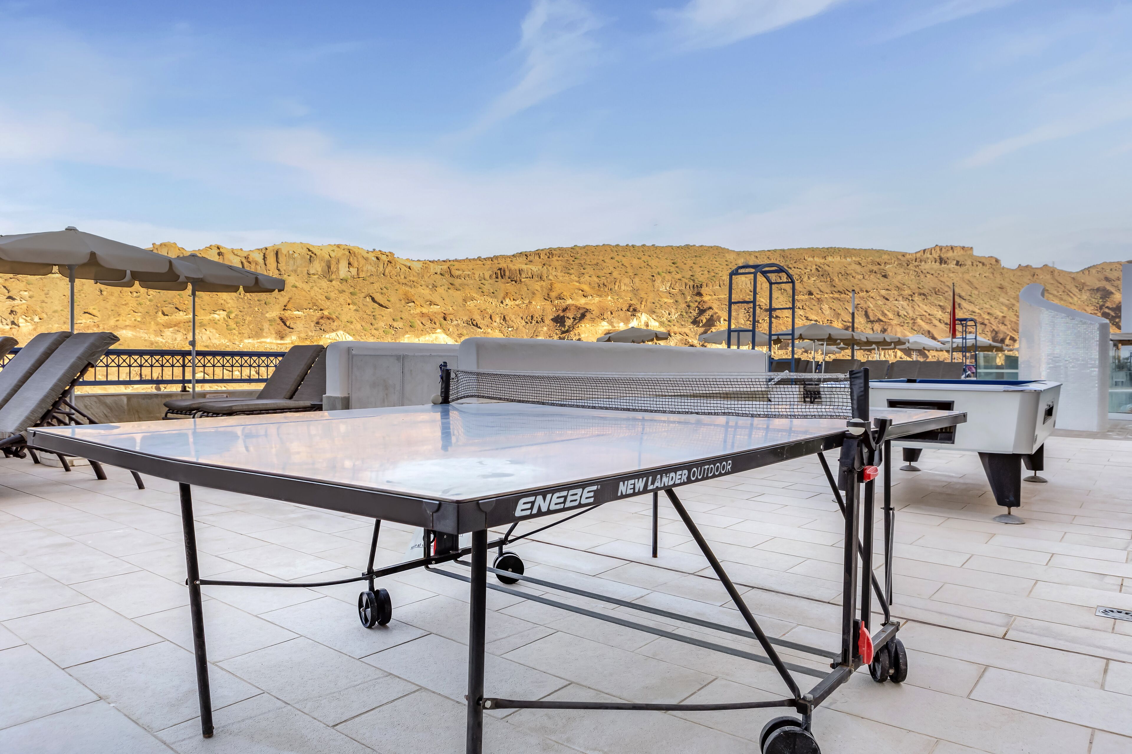 Mesa de ping pong con red, instalada en un patio con baldosas, sillas de descanso y sombrillas.