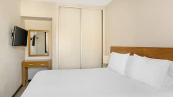 A hotel room with a bed, a TV mounted on the wall, a wooden nightstand, a mirror, and a wardrobe.