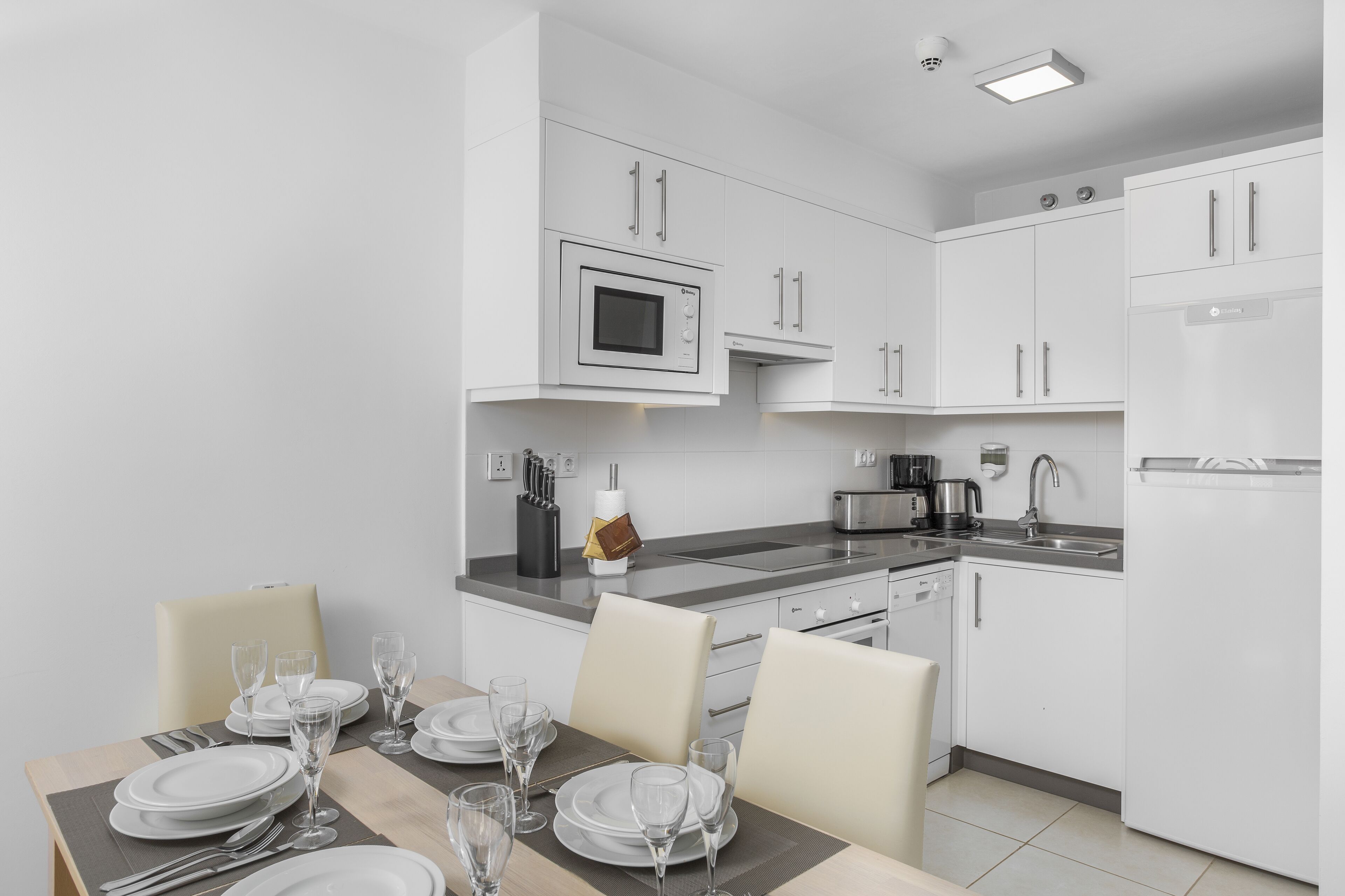 A modern kitchen with white cabinetry, a built-in microwave, and a dining area set for four with a table, chairs, and glassware.