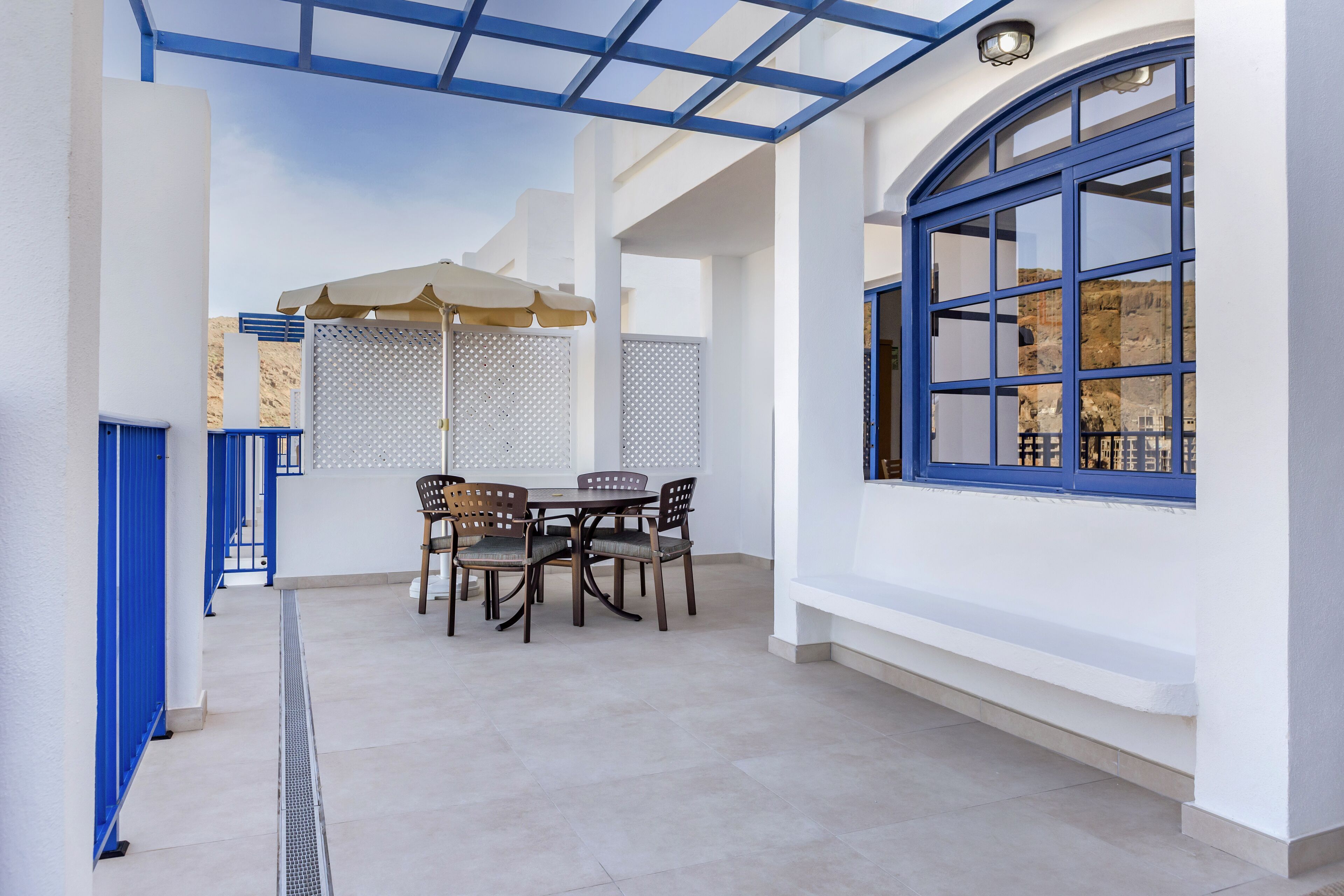 Un amplio patio con mesa y sillas, una pérgola con vigas azules y un ventanal con marco azul.