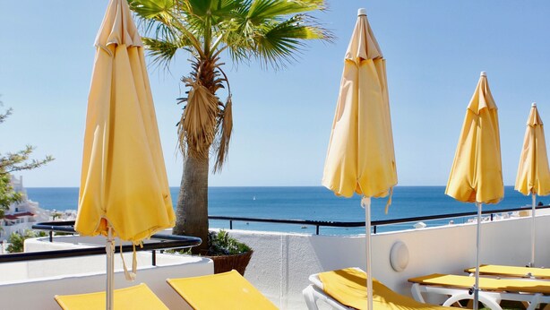 A terrace with yellow lounge chairs and umbrellas, a palm tree, and a view of the sea. - Alfagar Cerro Malpique (Albufeira)