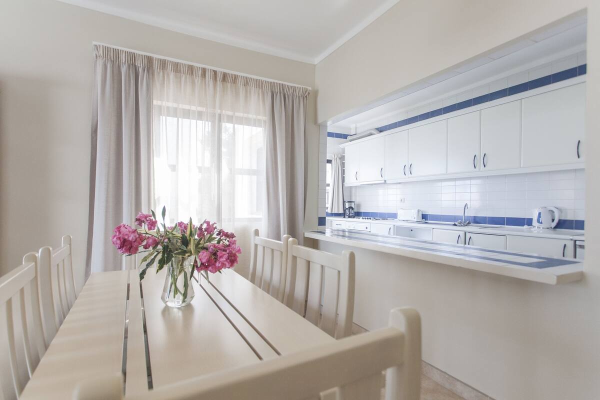 A dining area with a table set for four, a vase with flowers, and a kitchen with white cabinets and blue accents.