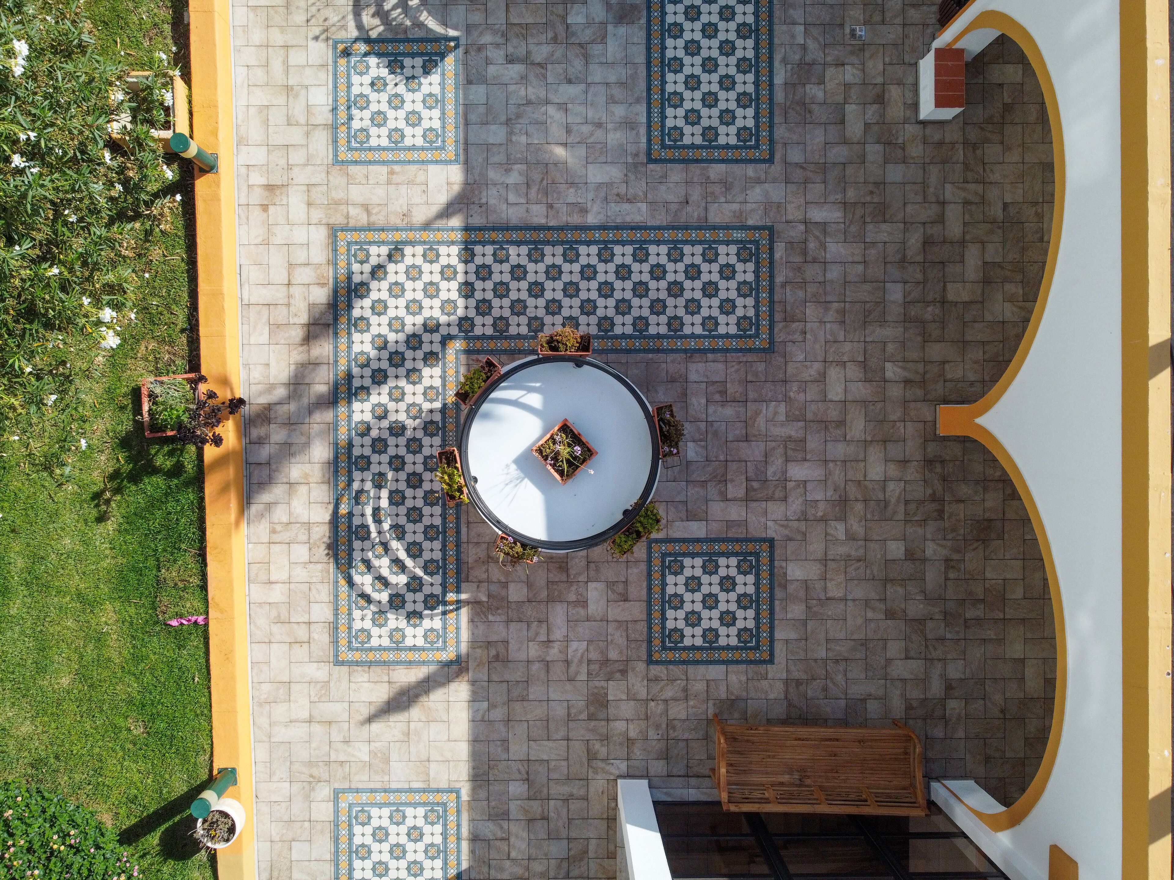 Overzicht vanuit de lucht van een binnenplaats met een ronde tafel in het midden, omringd door vierkante tegels met een geometrisch patroon, een bank en plantenbakken.