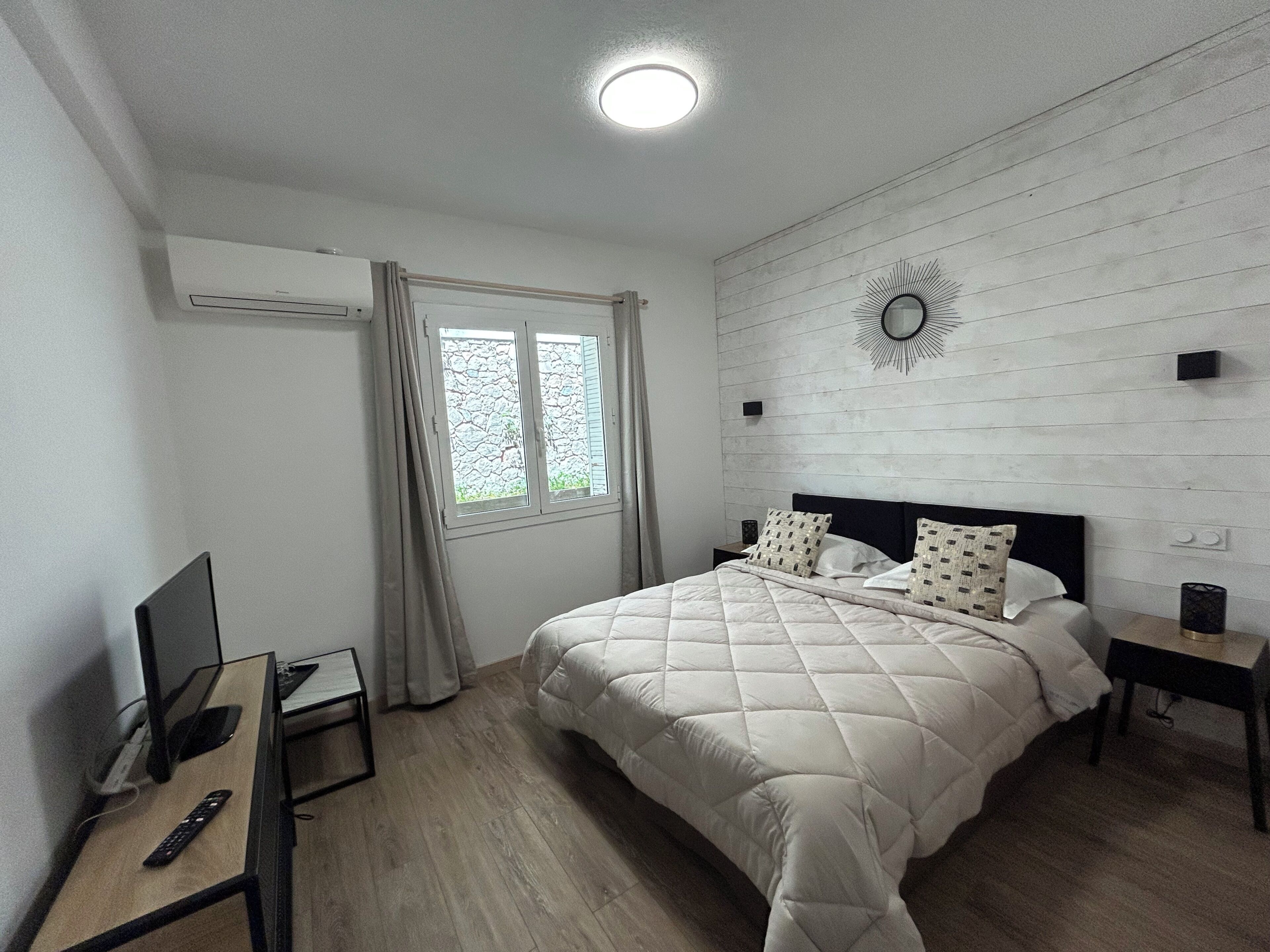 Un dormitorio con cama, un escritorio con computadora, un televisor, una ventana con cortinas, y una lámpara de pared.