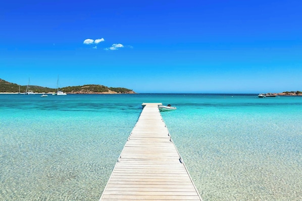 Un muelle de madera que se adentra en un mar azul y transparente, con barcos fondeados cerca.