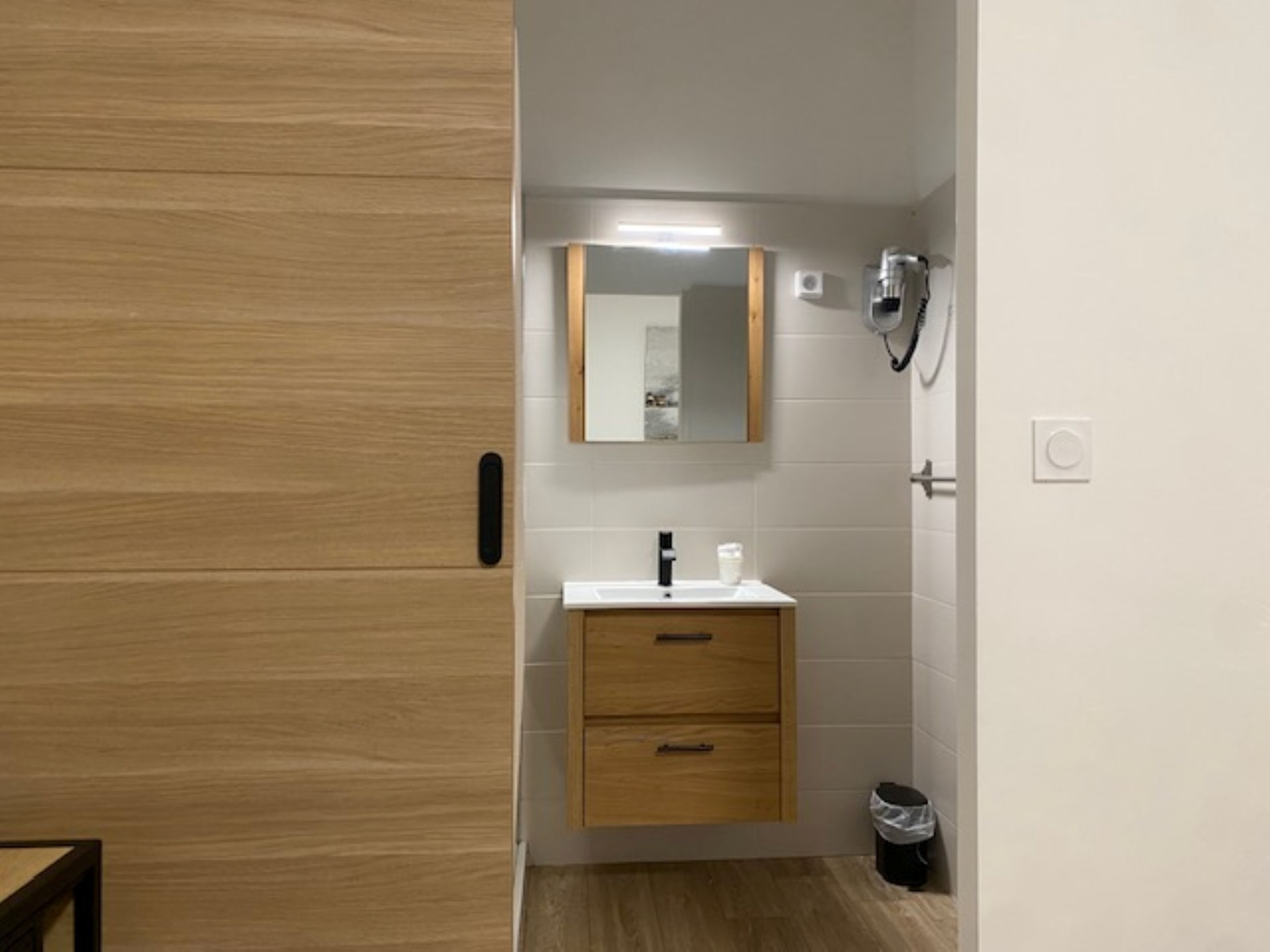Une salle de bain moderne avec une porte en bois, un miroir au-dessus d’un lavabo, une douchette à main et une poubelle.