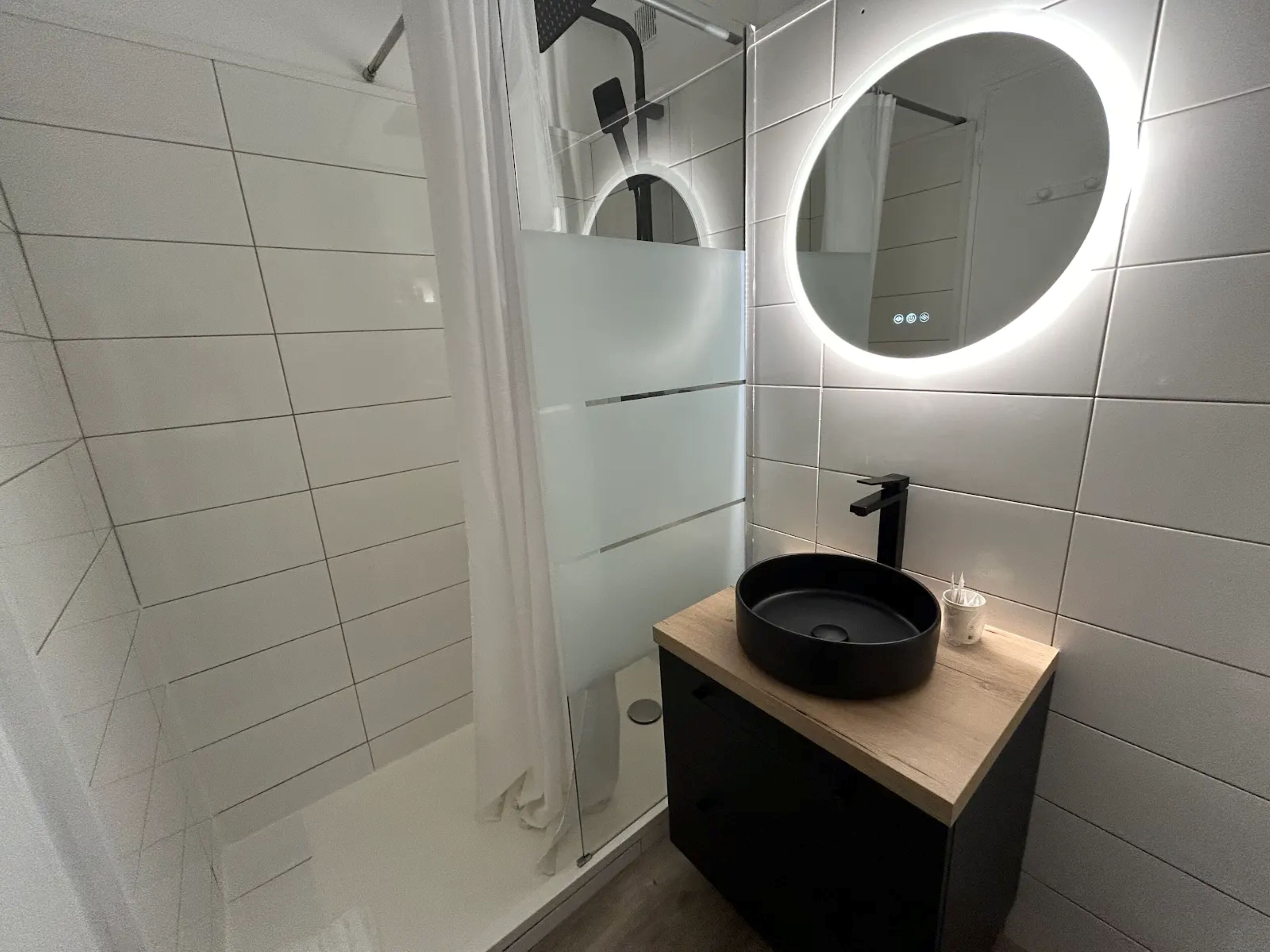 Un bagno moderno con cabina doccia in vetro, uno specchio rotondo con illuminazione integrata e un lavandino nero su un piano in legno.
