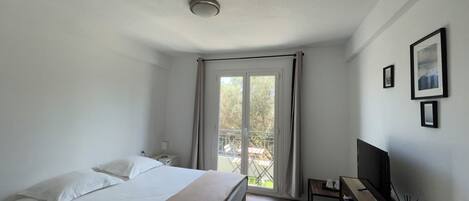 A hotel room with a bed, a desk with a TV, a window with curtains, and a balcony.