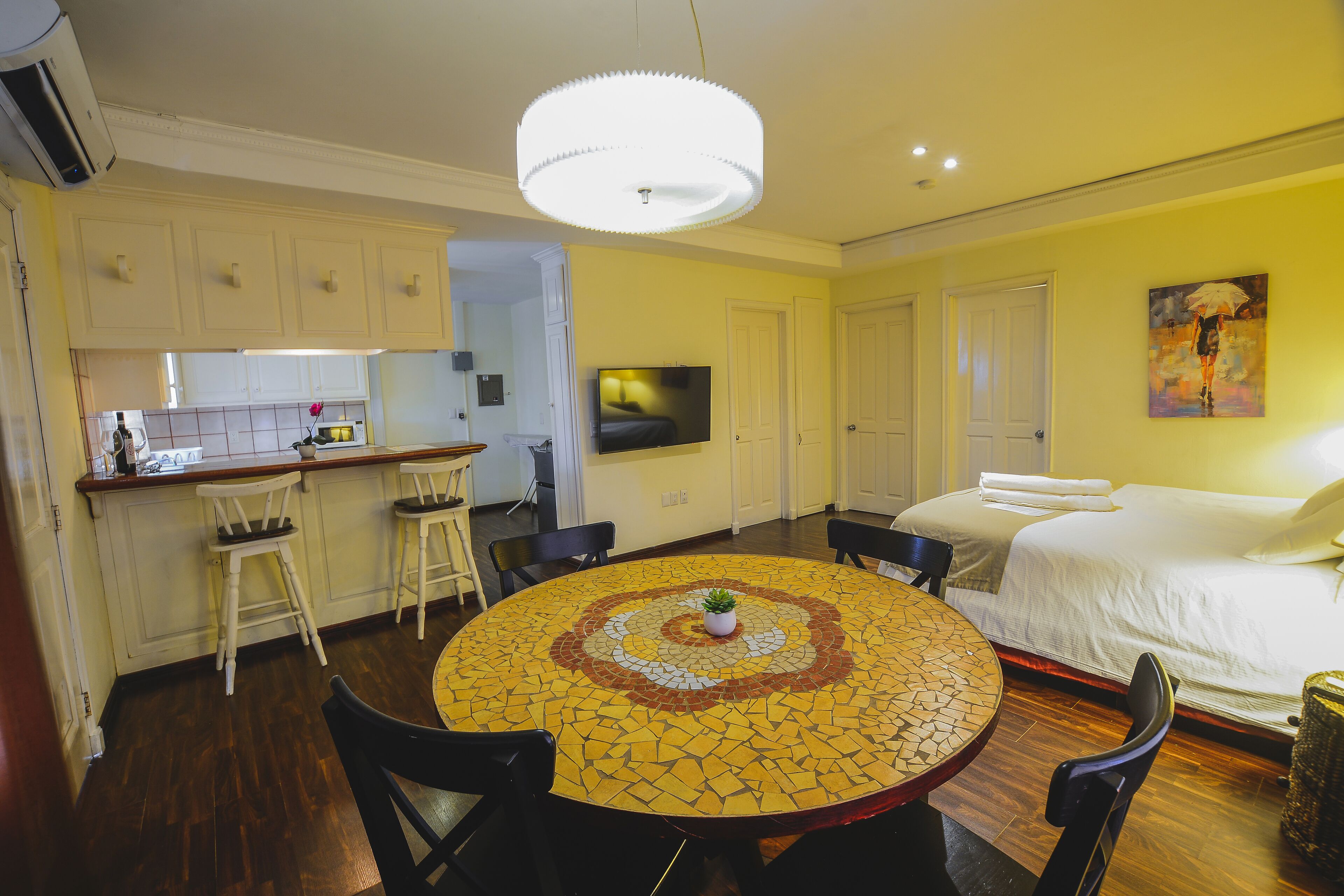 A hotel room with a bed, a dining table with chairs, a kitchen area, and a TV.