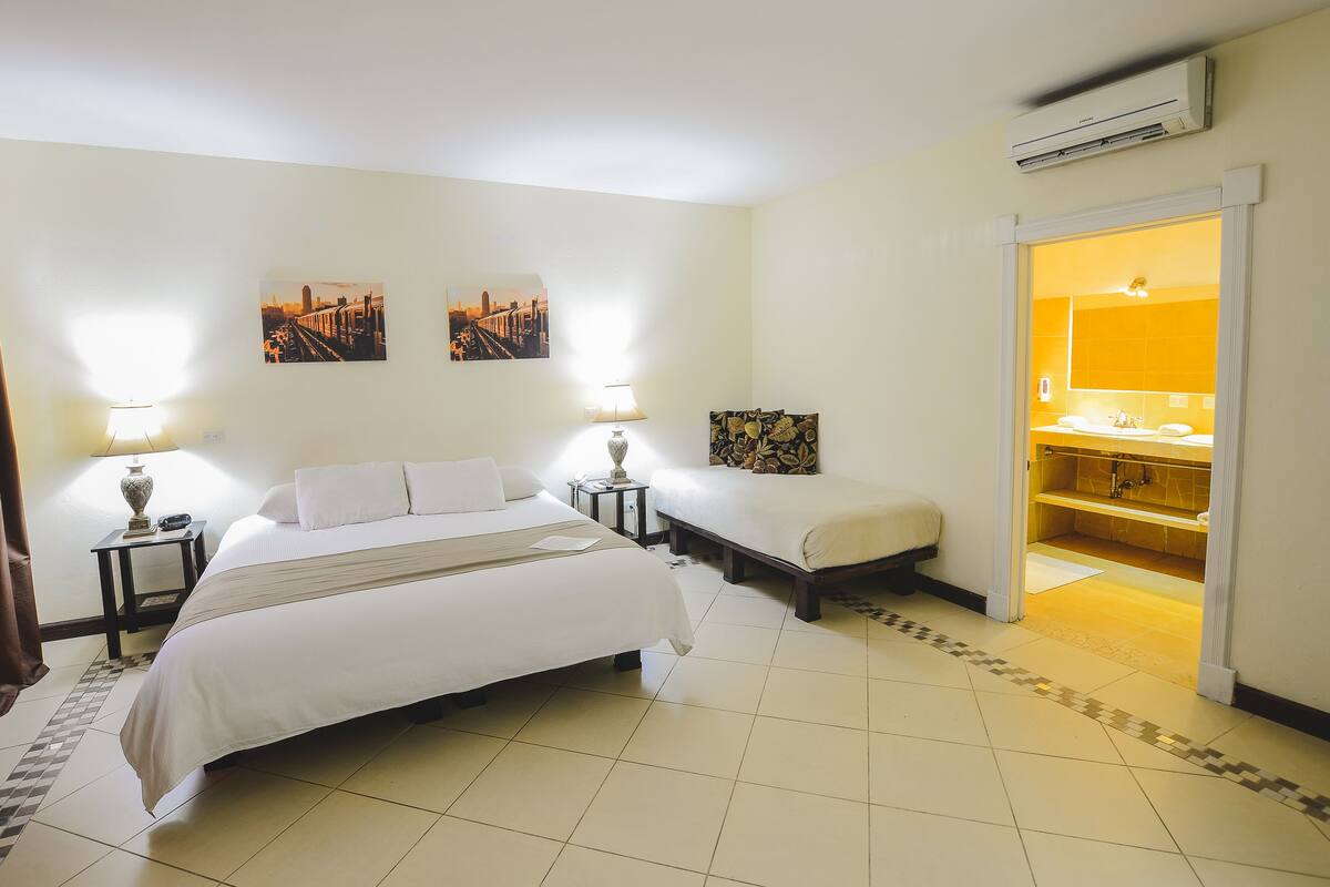 Ein Hotelzimmer mit einem großen Bett, einem kleineren Bett, einem Nachttisch mit Lampe, einem Bild an der Wand und einer offenen Badezimmertür.