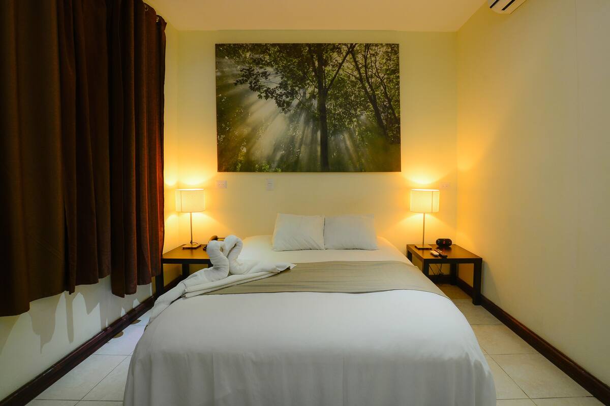 Ein Hotelzimmer mit einem großen Bett, zwei Nachttischlampen, einem Waldgemälde und einem braunen Vorhang.