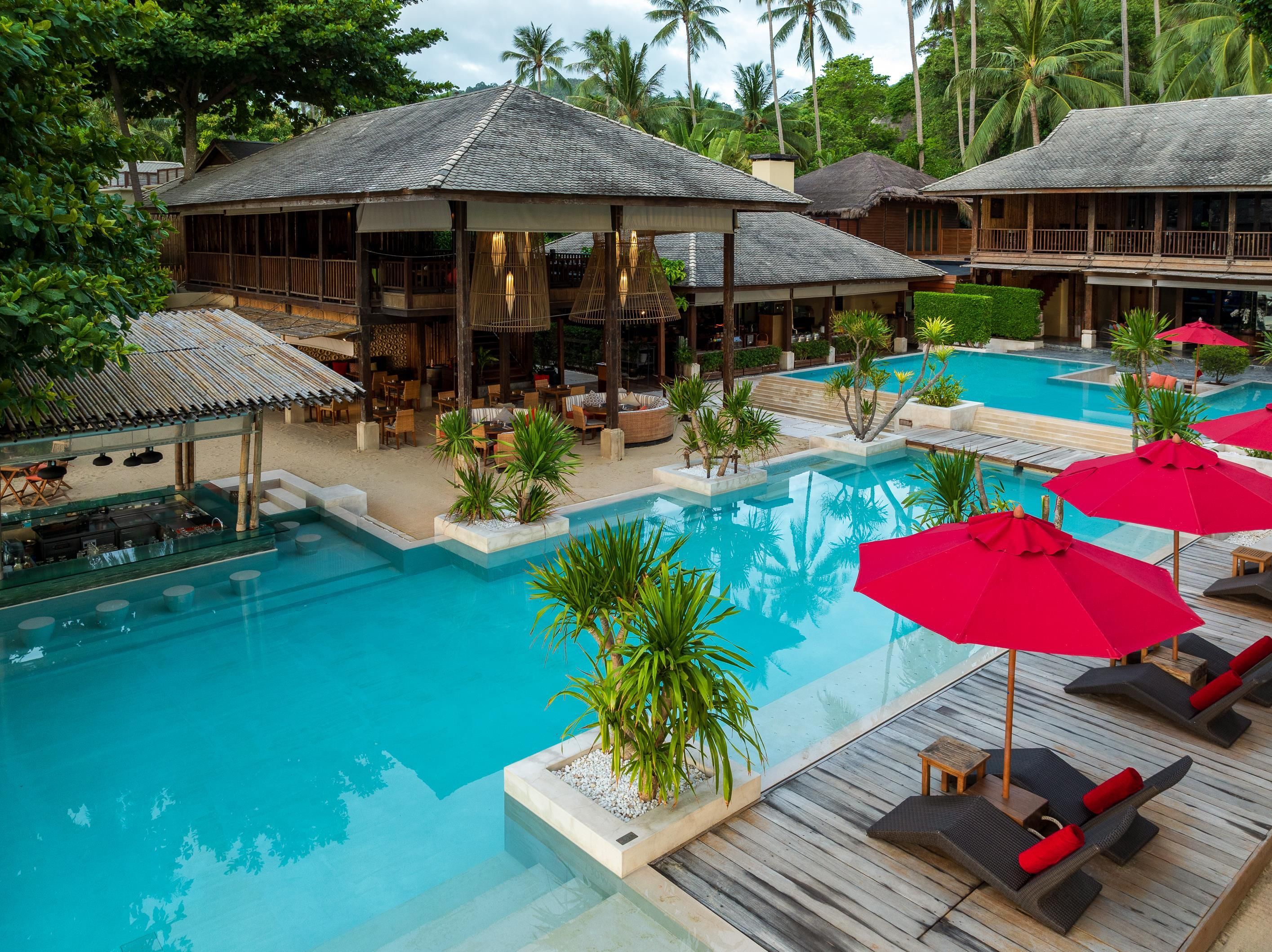 Un espace piscine aménagé dans un complexe hôtelier, avec de nombreux chaises longues et parasols, entouré de structures en bois et d’une végétation tropicale.