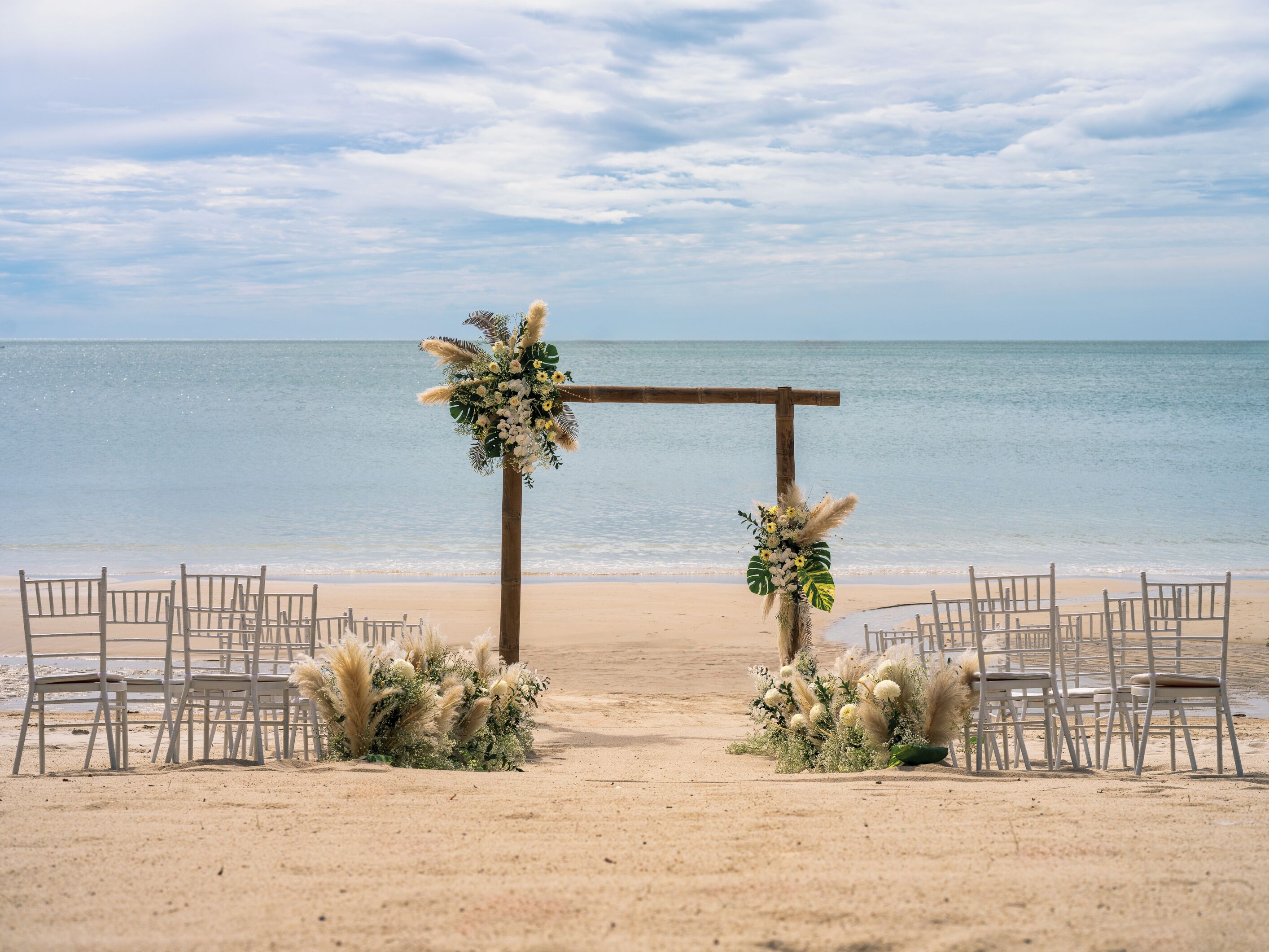 A beach wedding setup with a wooden arch adorned with flowers and pampas grass, surrounded by white chairs.