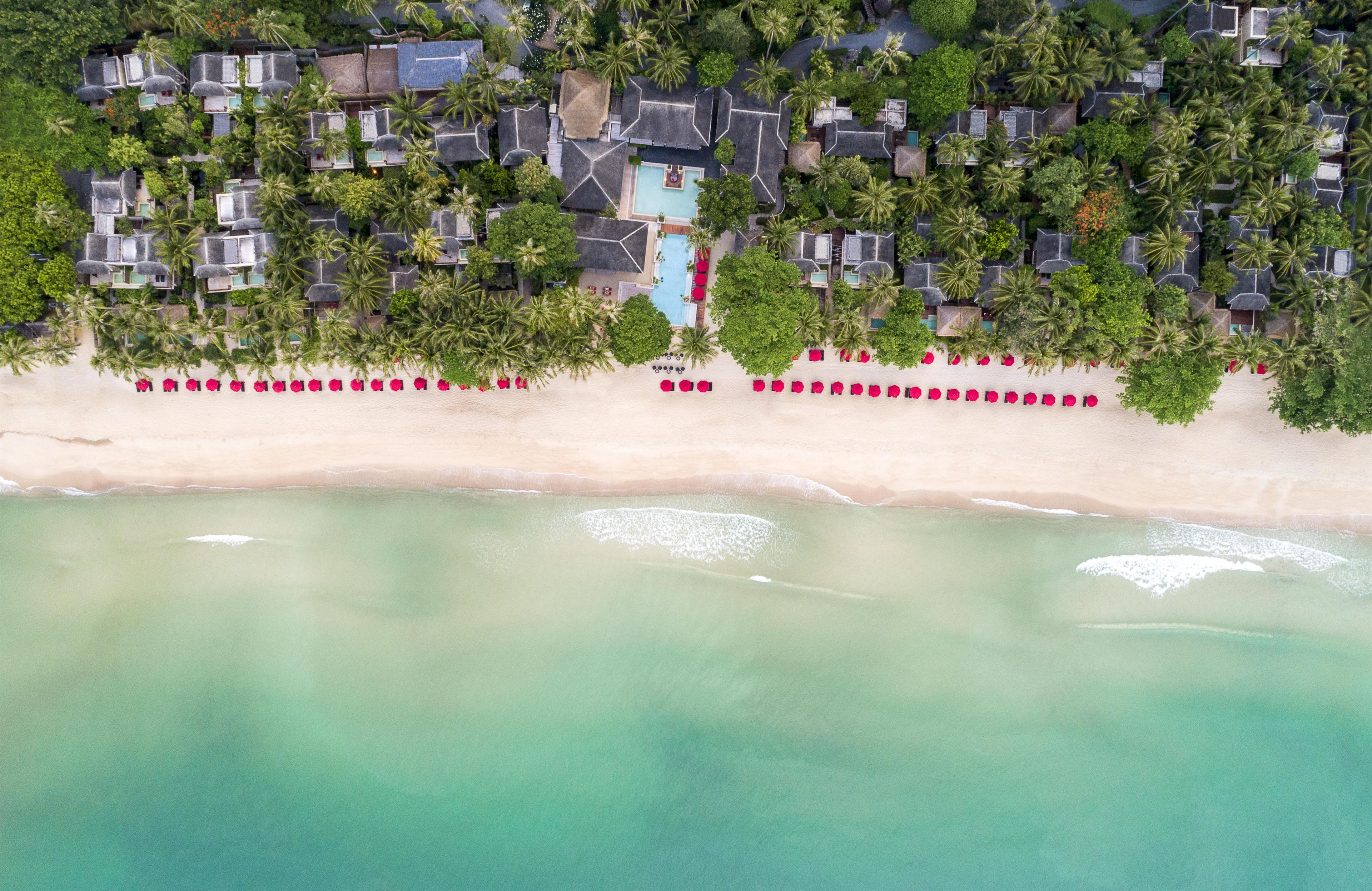 An aerial view of a beachfront resort with multiple bungalow-style accommodations, a swimming pool, and a sandy beach.