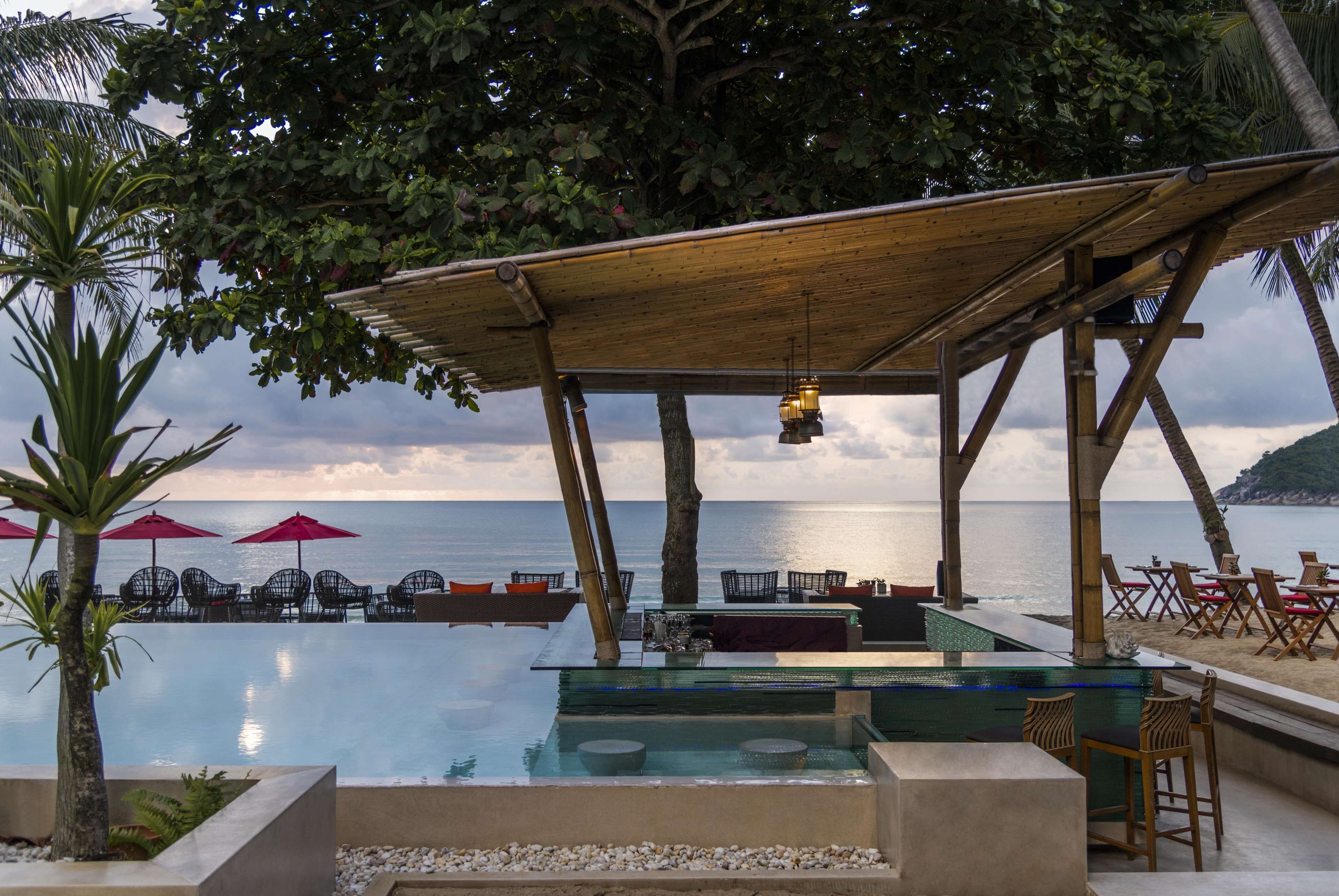 An infinity pool with a wooden pergola overhead, surrounded by lounge chairs and umbrellas, overlooking the sea.
