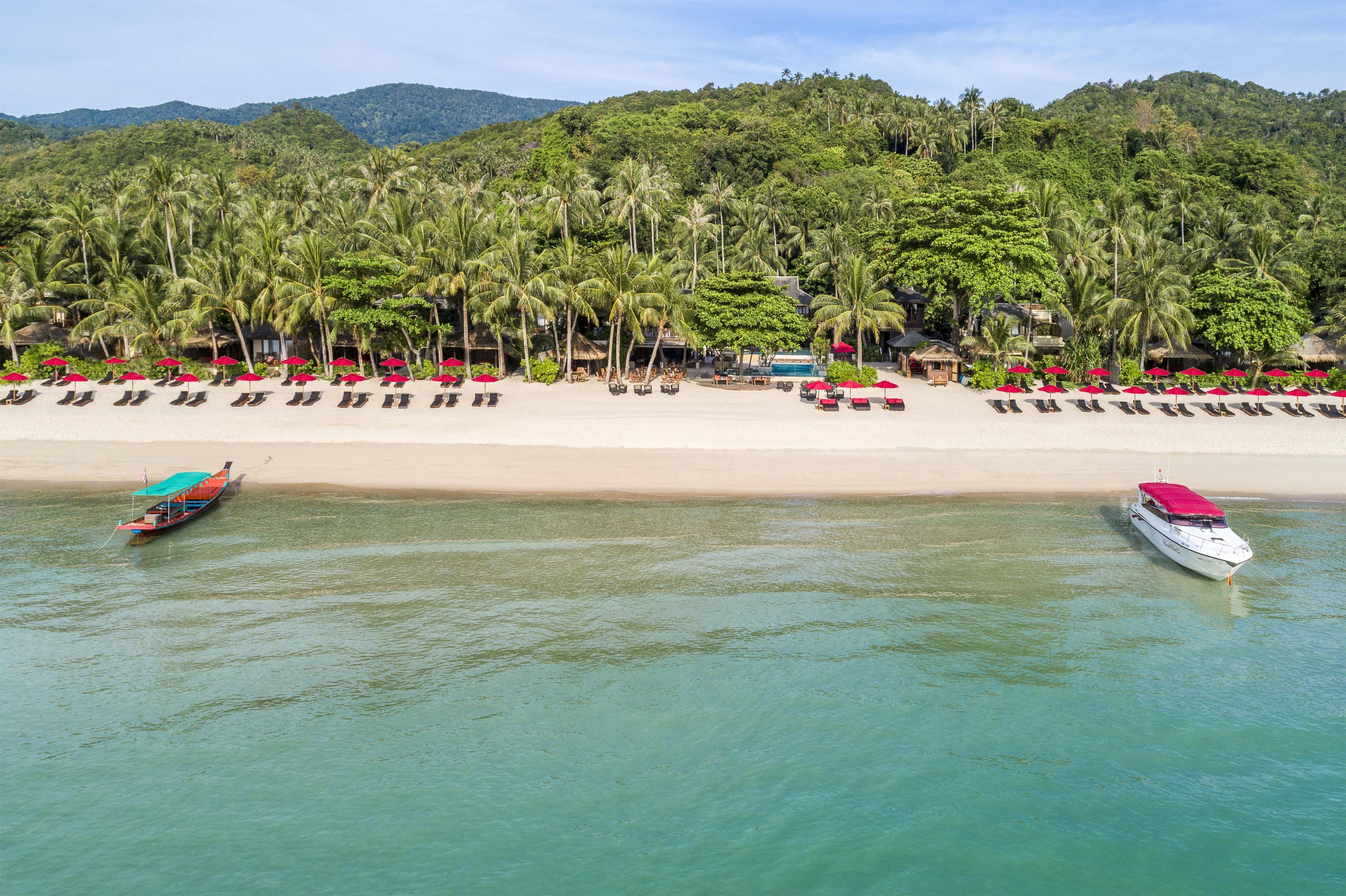 Une plage bordée de rangées de chaises longues et de parasols, une piscine et des bateaux sur l’eau.